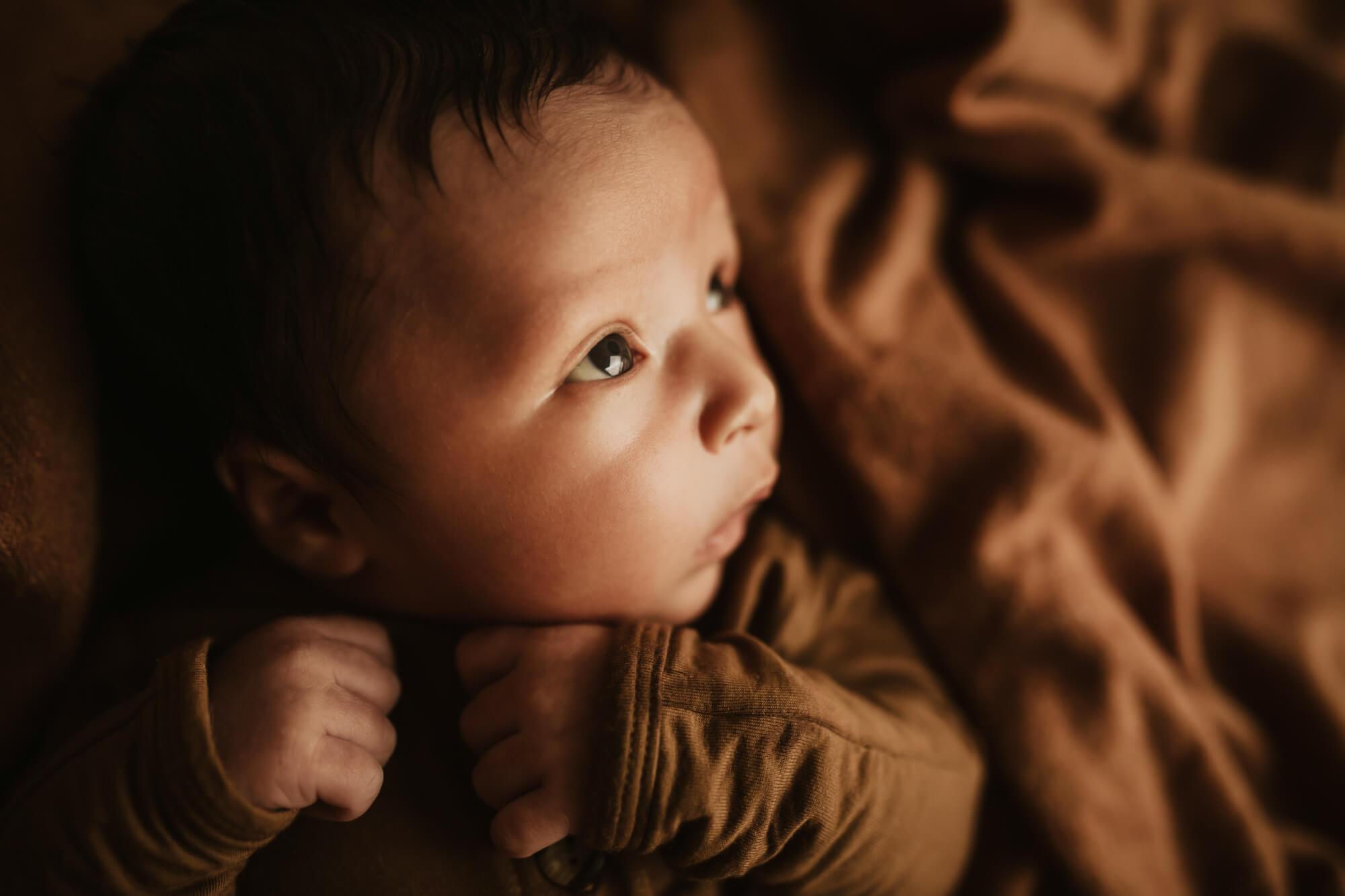 A newborn baby lays on a brown blanket while looking up at a window