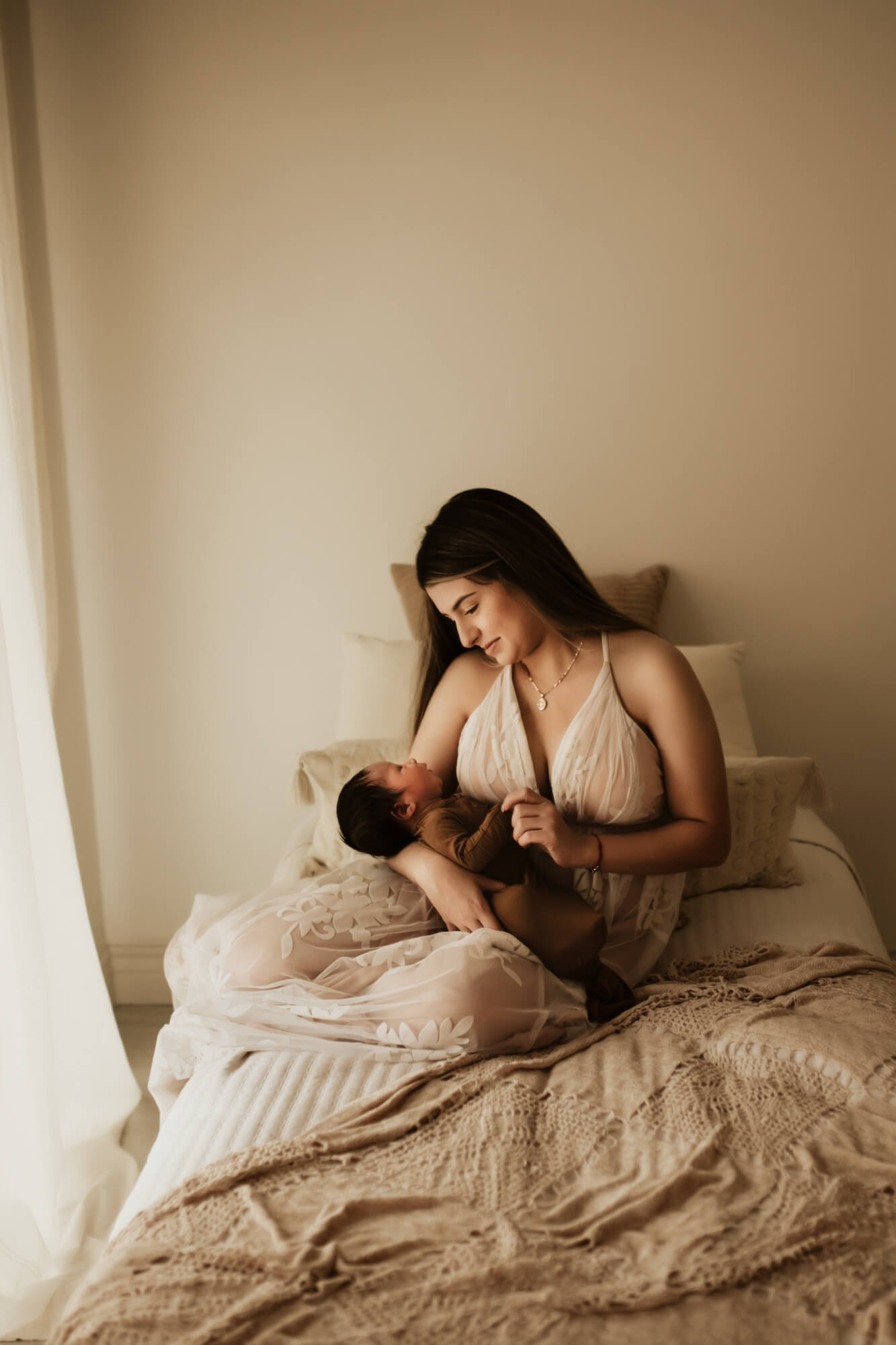 A new mother in a white lace gown sits on a bed holding her newborn baby by a window beautifully connected