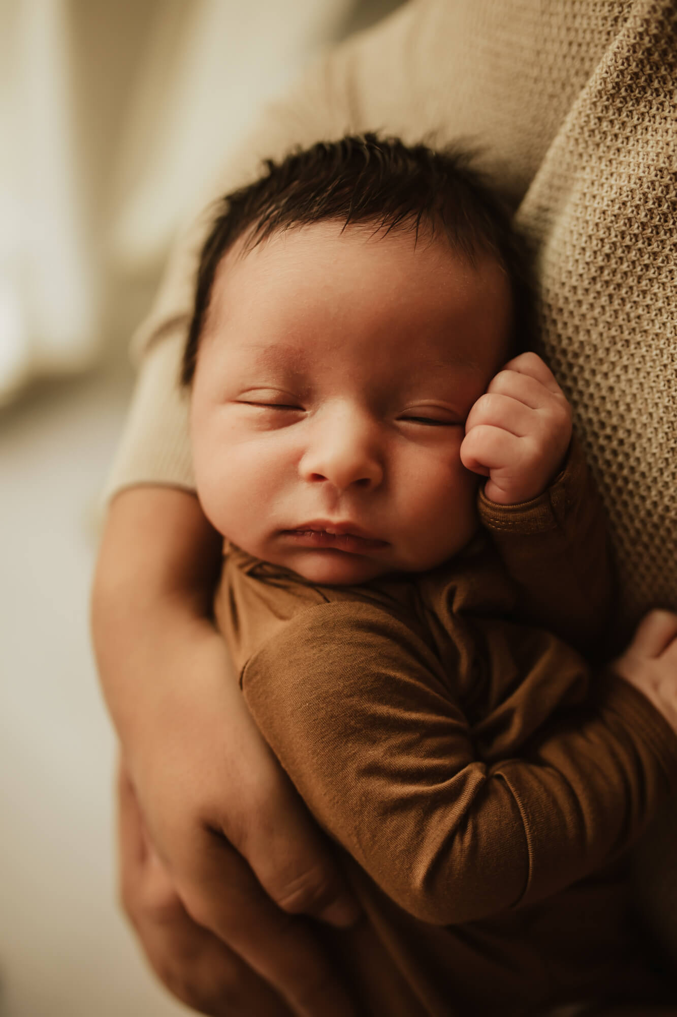 A newborn baby sleeps against mom's chest wearing a brown onesie beautifully-connected