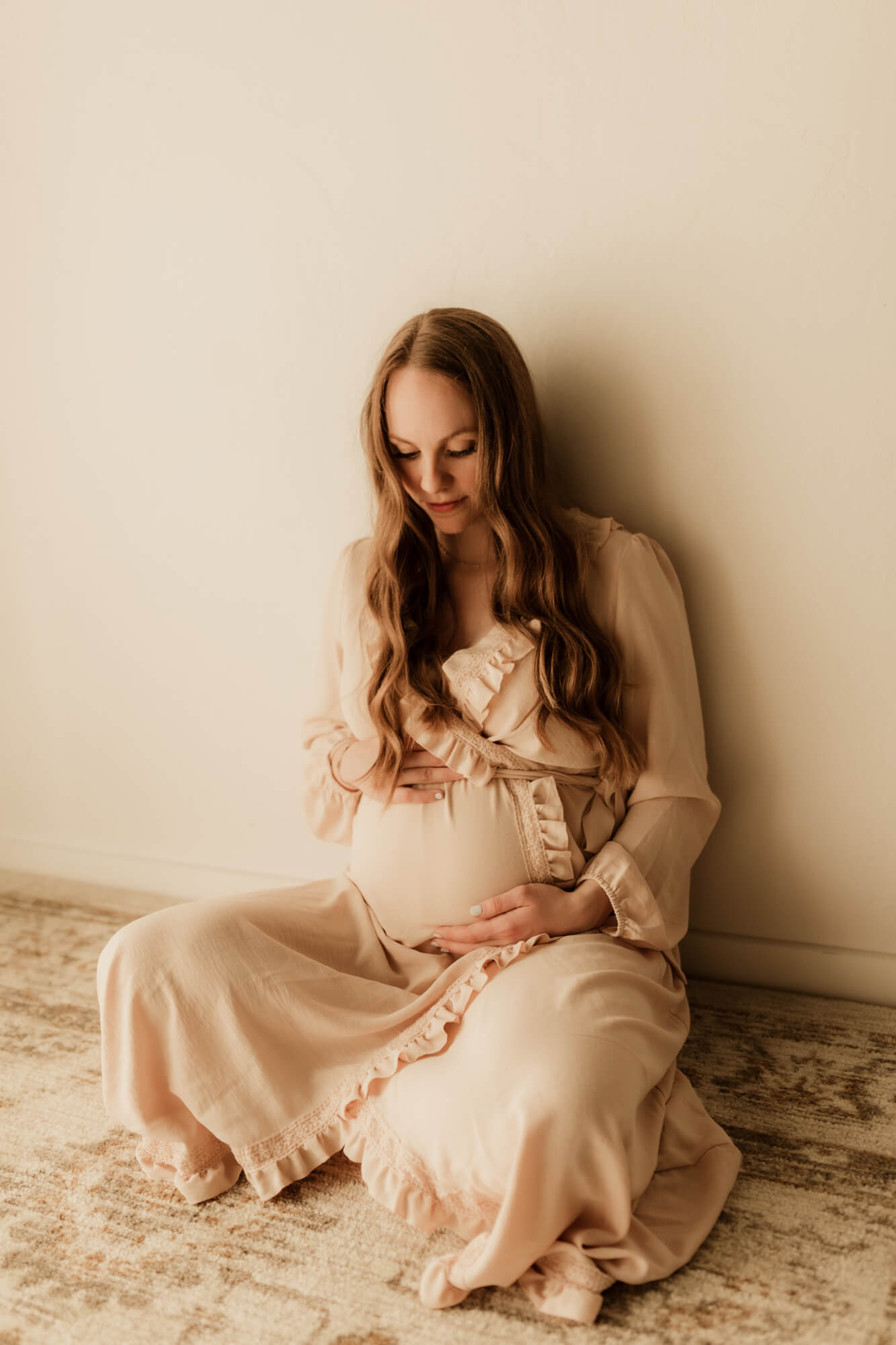 A mom-to-be sits on the floor against a wall holding and looking down on her bump midwife oklahoma city
