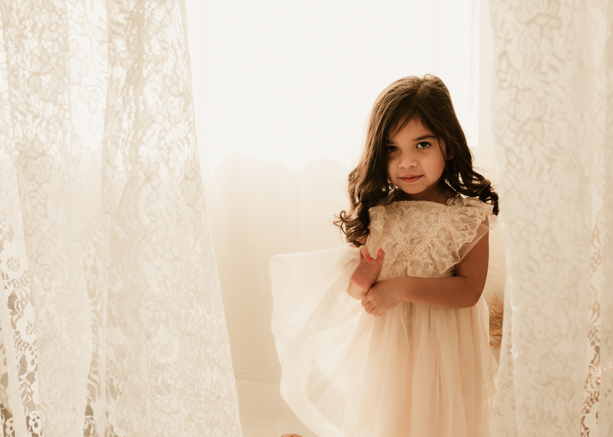A young girl plays with her lace and tule dress amongst some window curtains pediatrician OKC