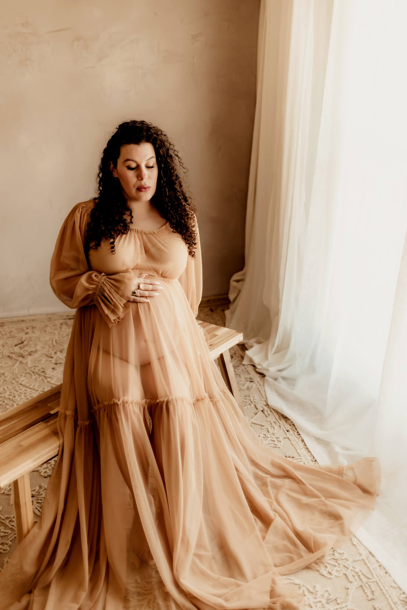 Woman with black curly hair sitting on a bench wearing a sheer tan dress and looking at her belly, beautifully connected.
