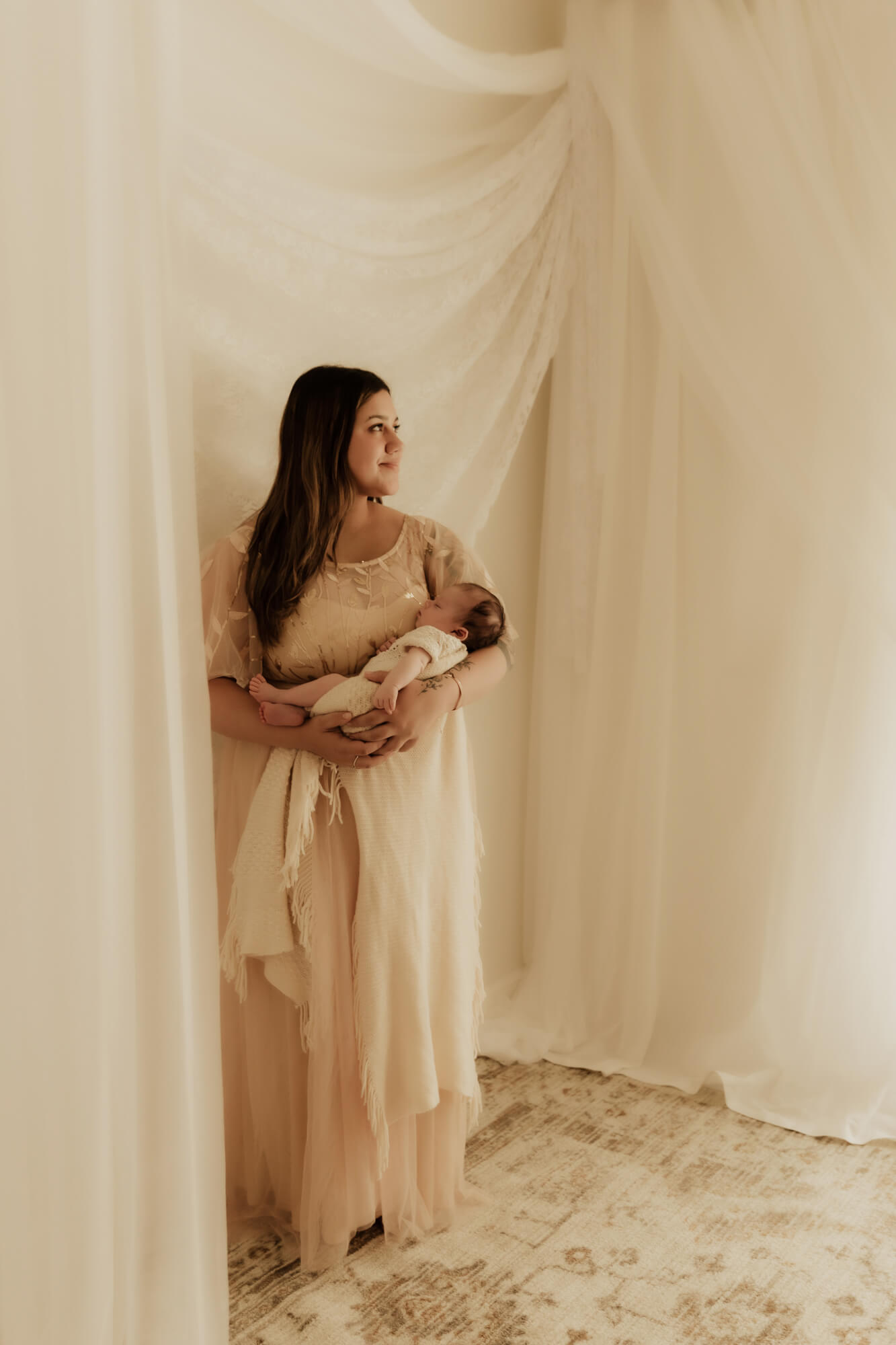 A mother stands in a corner of a room surrounded by drapes while holding her sleeping newborn baby