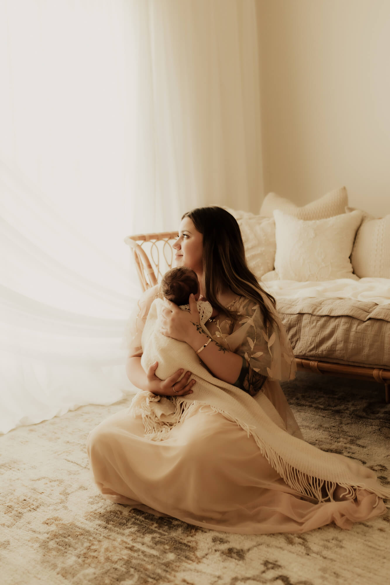 A new mom sits on the floor on a rug in front of a wicker couch by a window holding her newborn baby interior designer okc