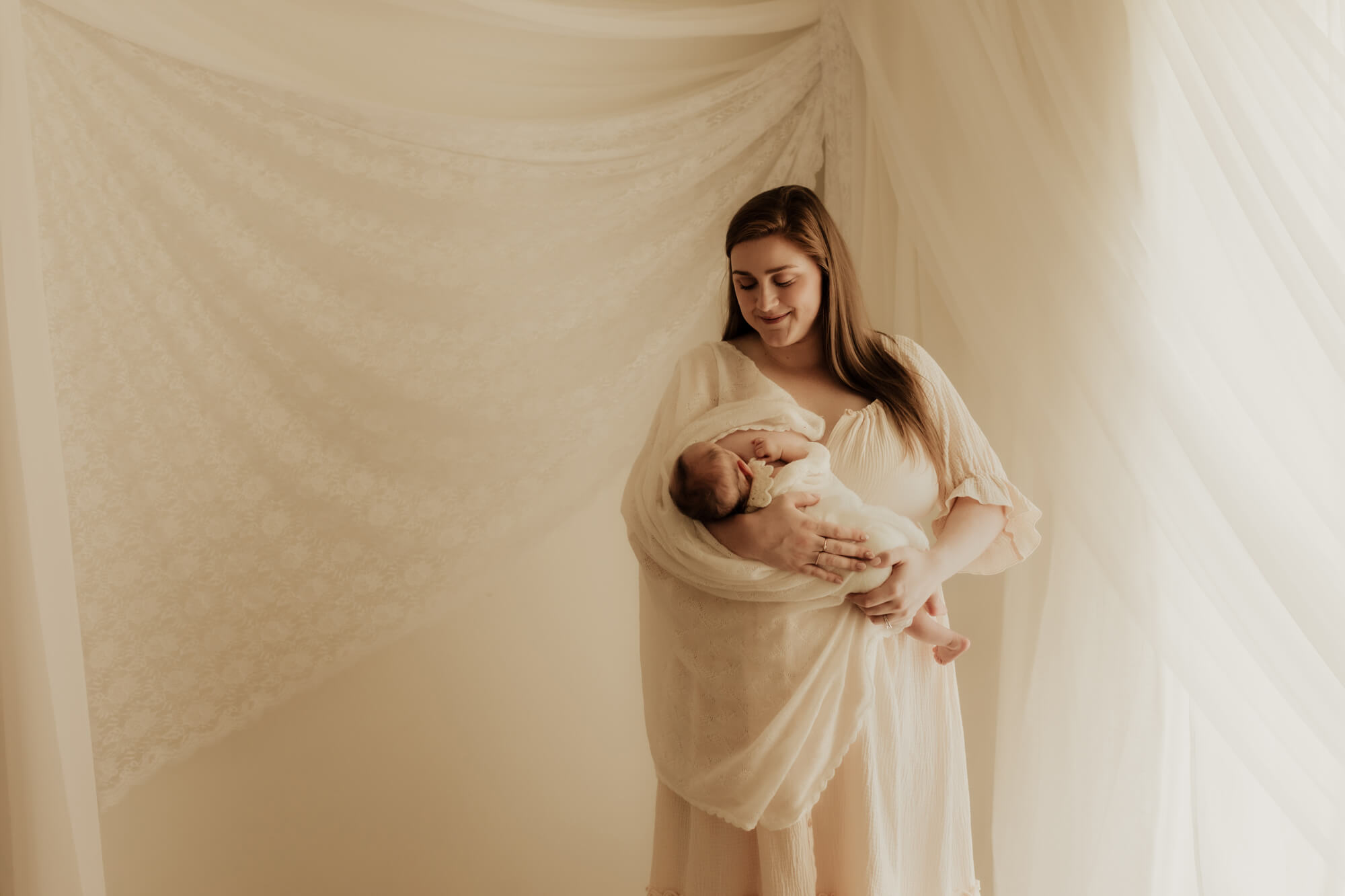 A mother stands in a studio in a white dress while breastfeeding her newborn daughter