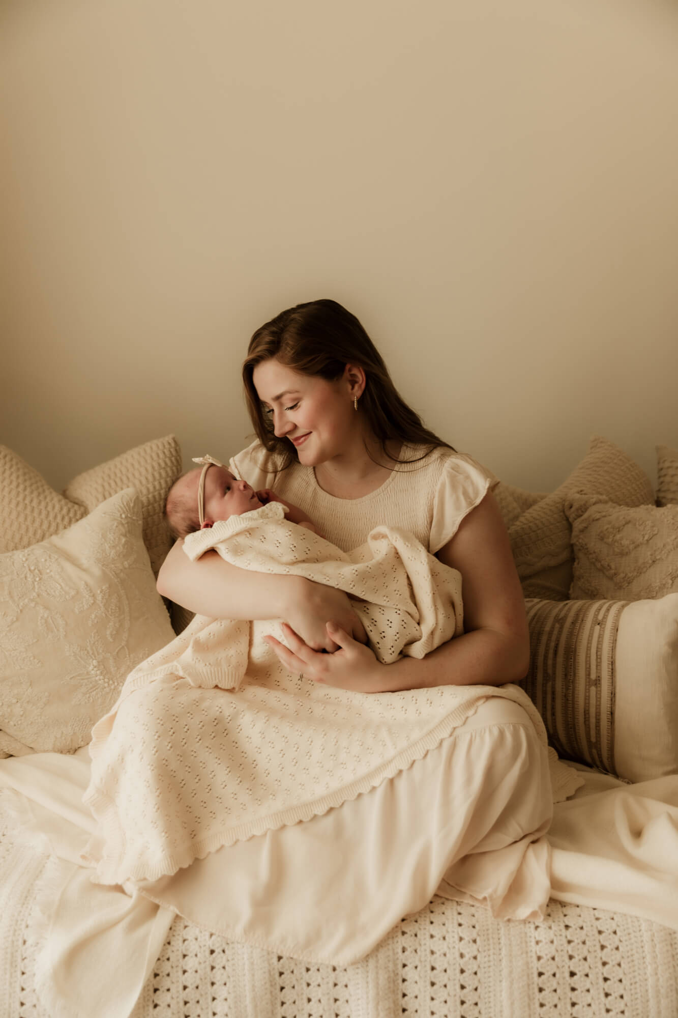 A mother in a white dress sits on a bed with pillows holding her newborn baby daughter in her arms lactation consultant okc