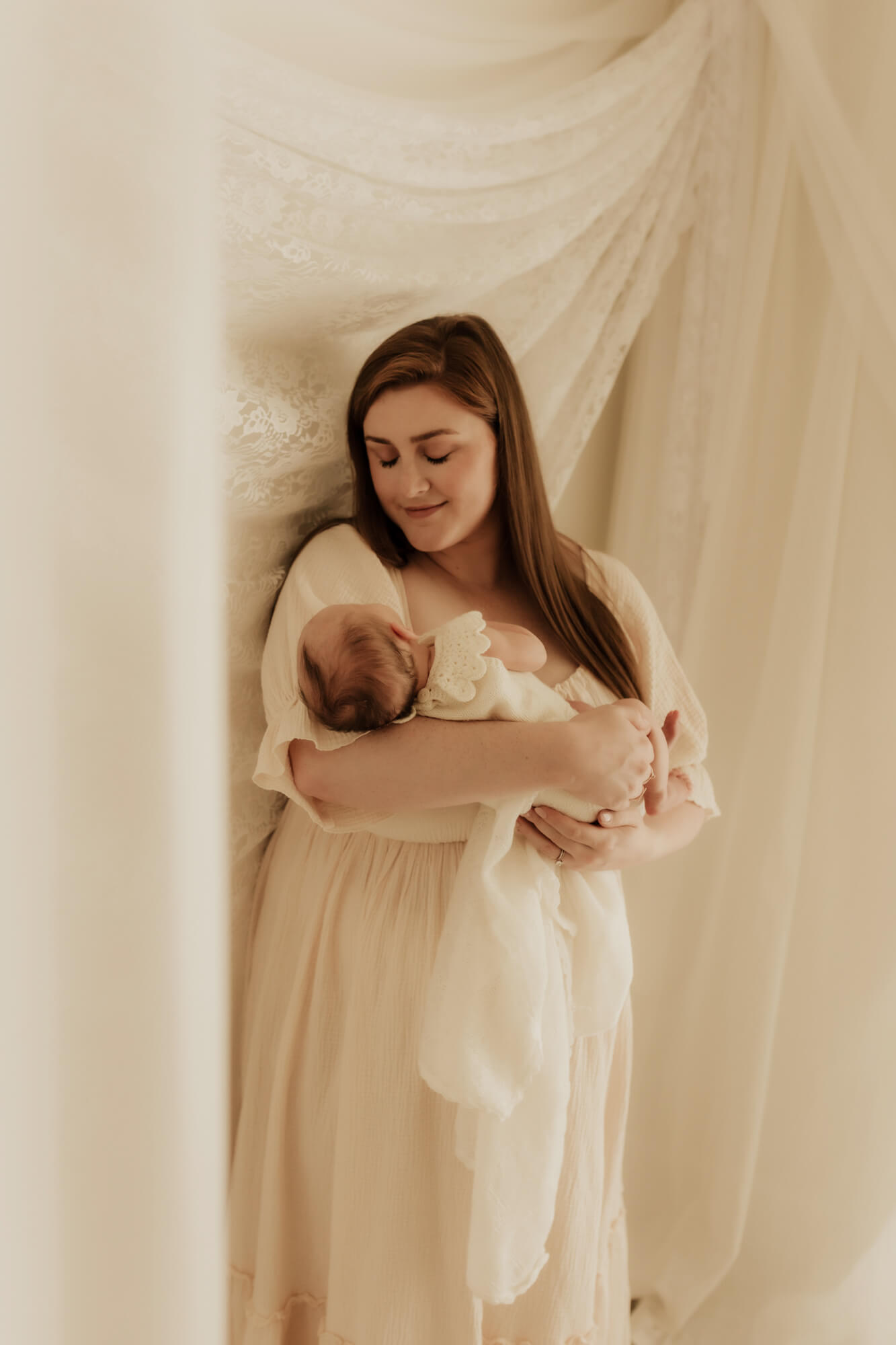 A new mother stands under some drapes looking down at the newborn baby in her arms lactation consultant okc