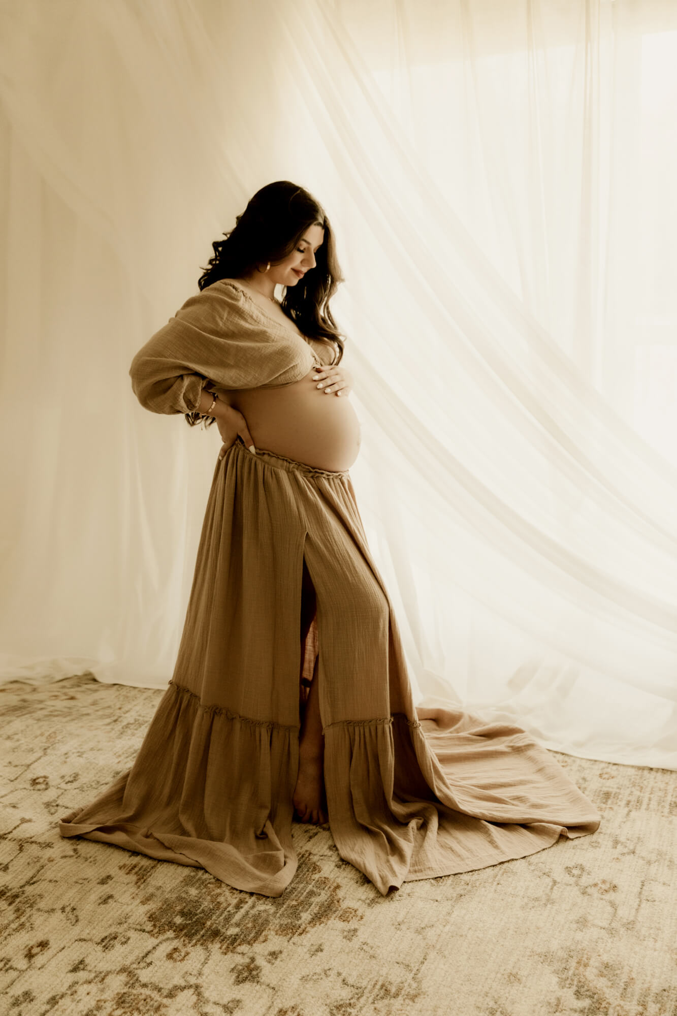 A mother to be stands in a studio by a window while looking down at her bump
