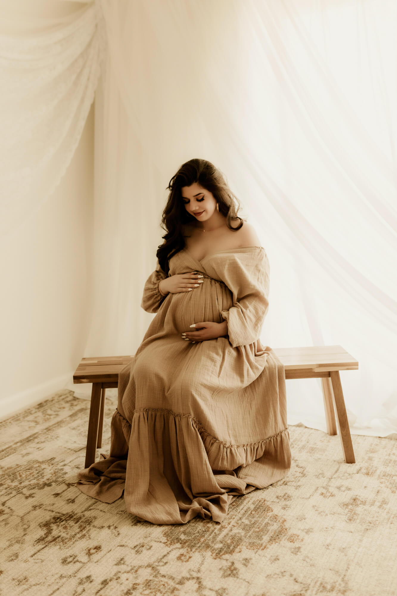 A mom to bee in a brown maternity gown sits on a wooden bench in a studio by a window OKC OBGYN