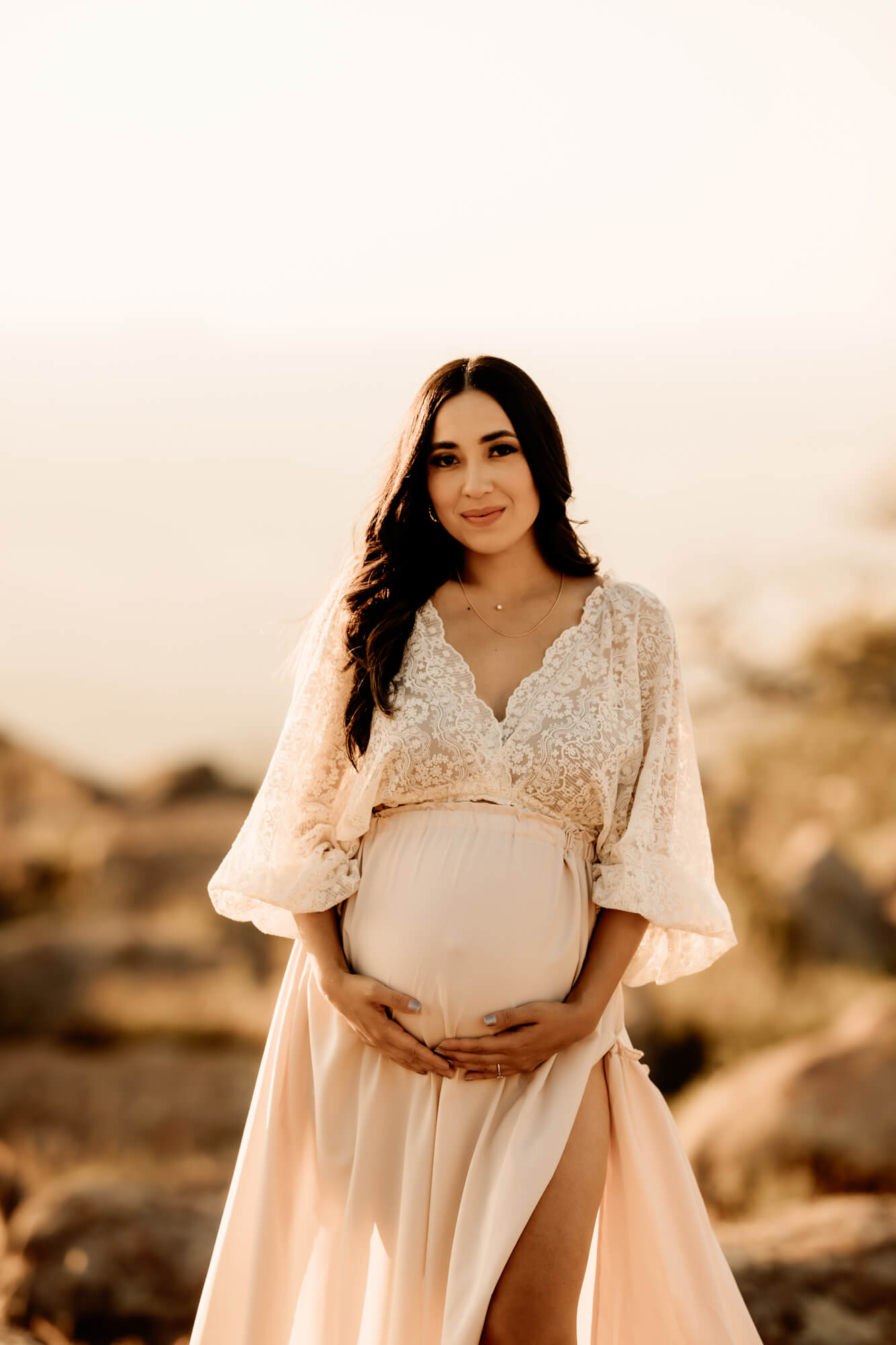 Expecting mother holds her baby bump while wearing a white dress, Modern Village Midwifery.