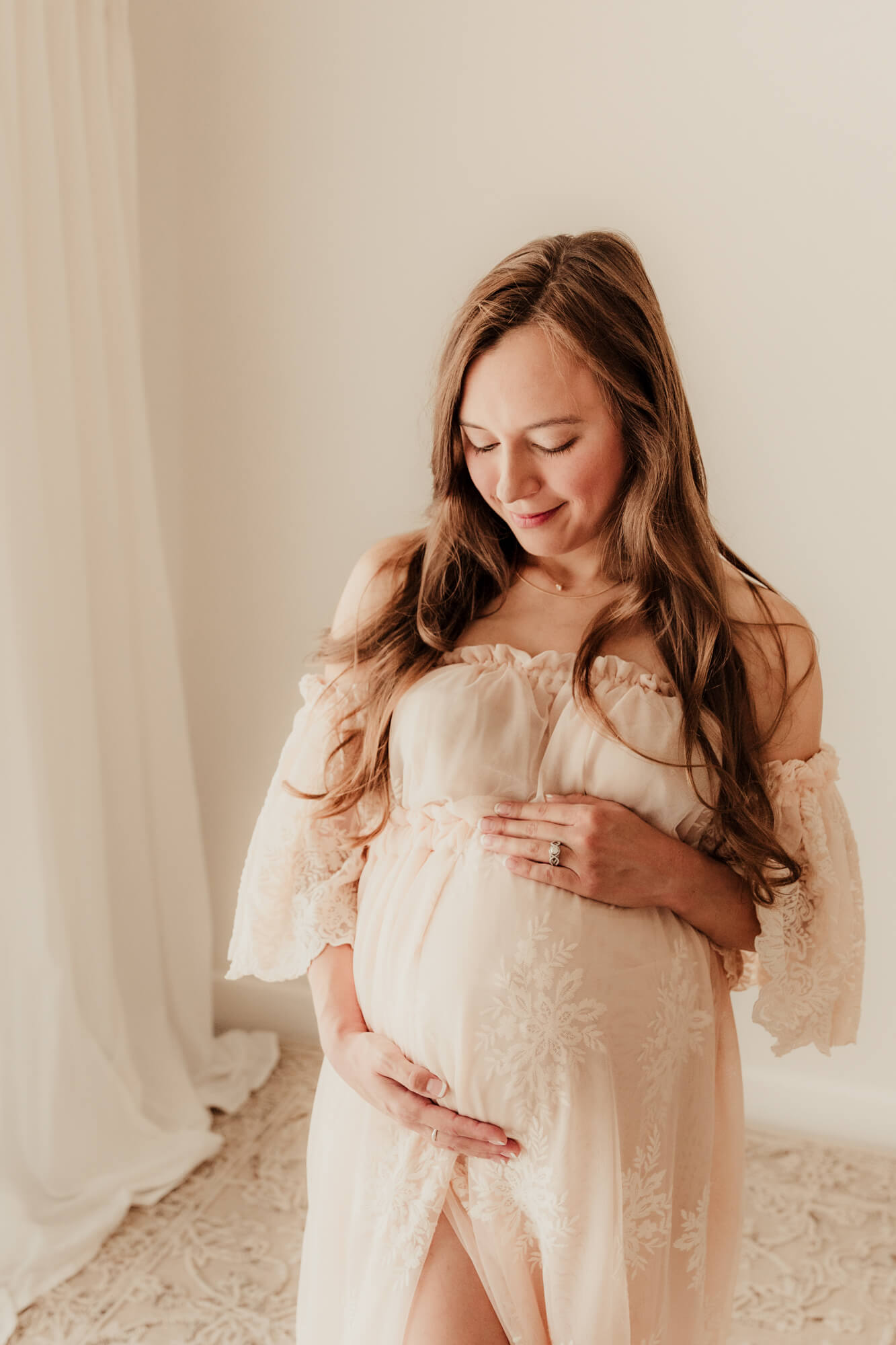 Woman looks at her baby bump while standing and wearing an off the shoulder lace dress.