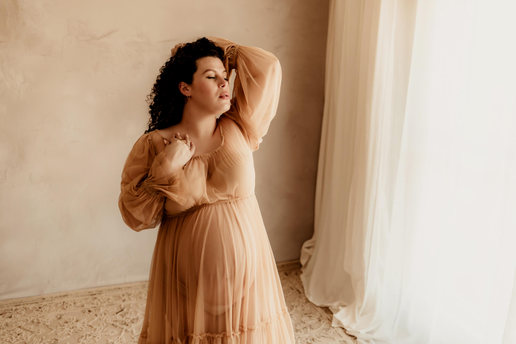 A pregnant woman in a sheer gown holds back her hair while standing in a window