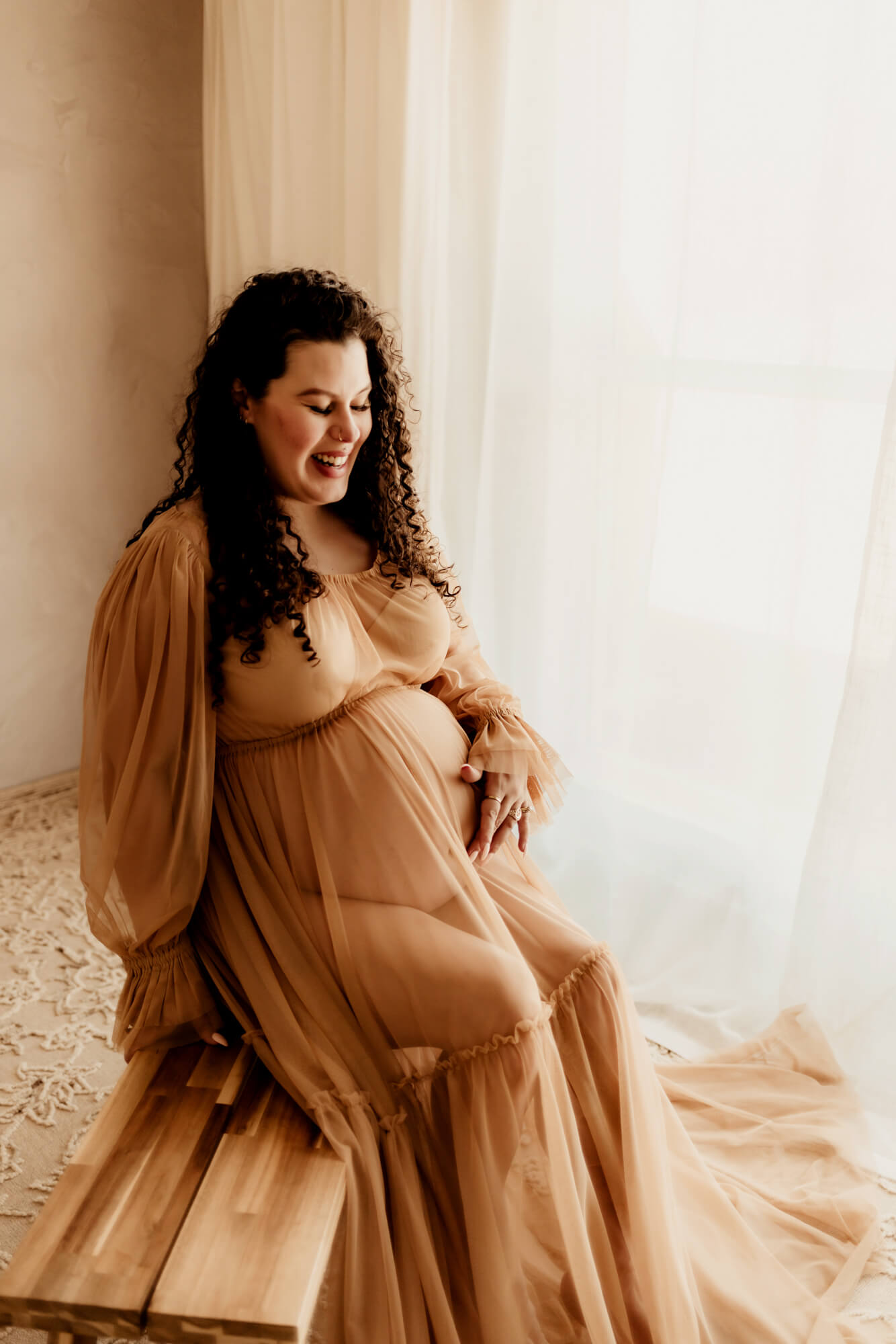 A mother to be smiles while sitting on a bench in a window in a brown sheer maternity gown thanks to Tulsa Fertility Center