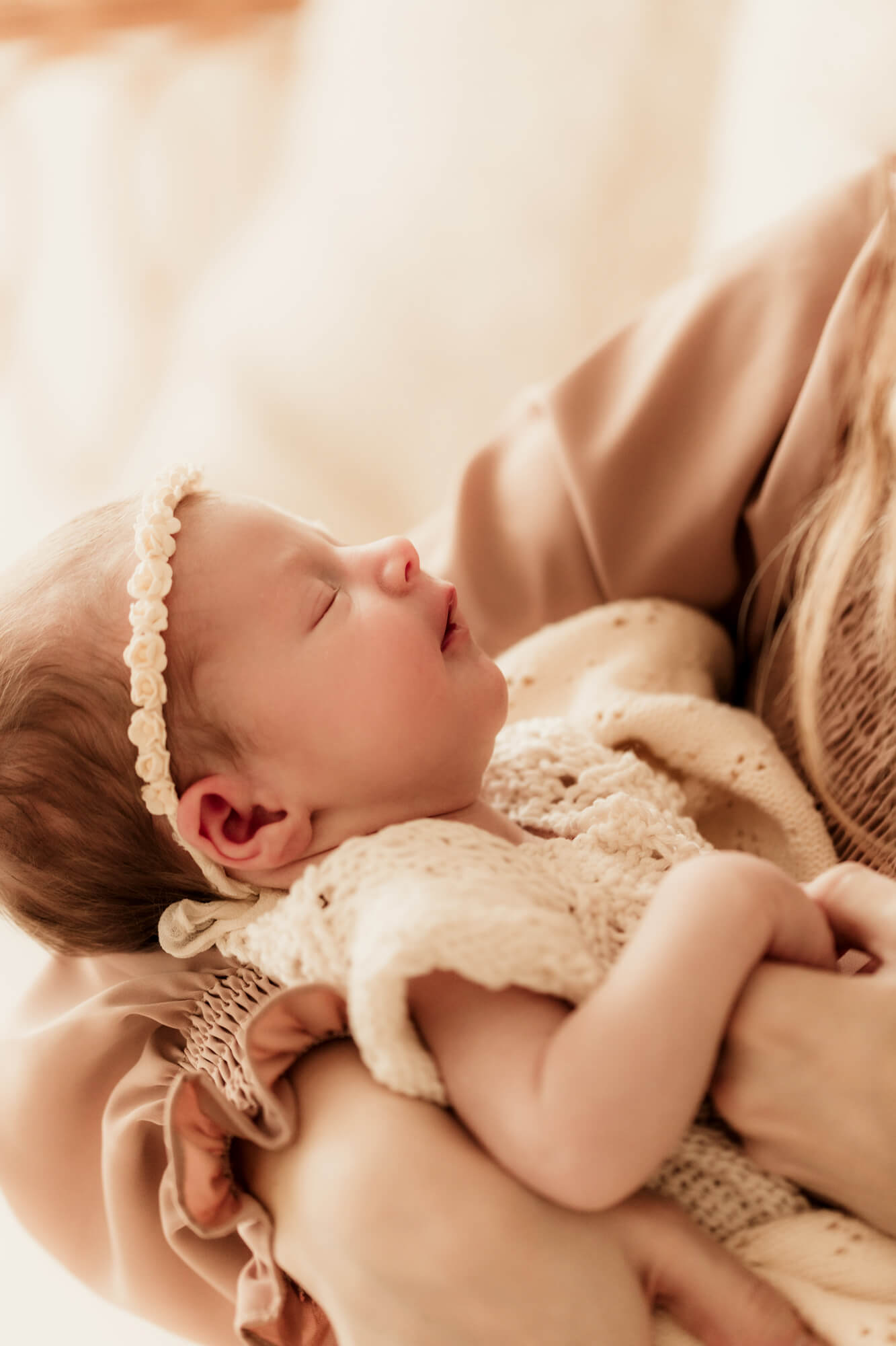 A baby girl in a white knit onesie and flower headband sleeps in mom's arms after some baby classes in OKC
