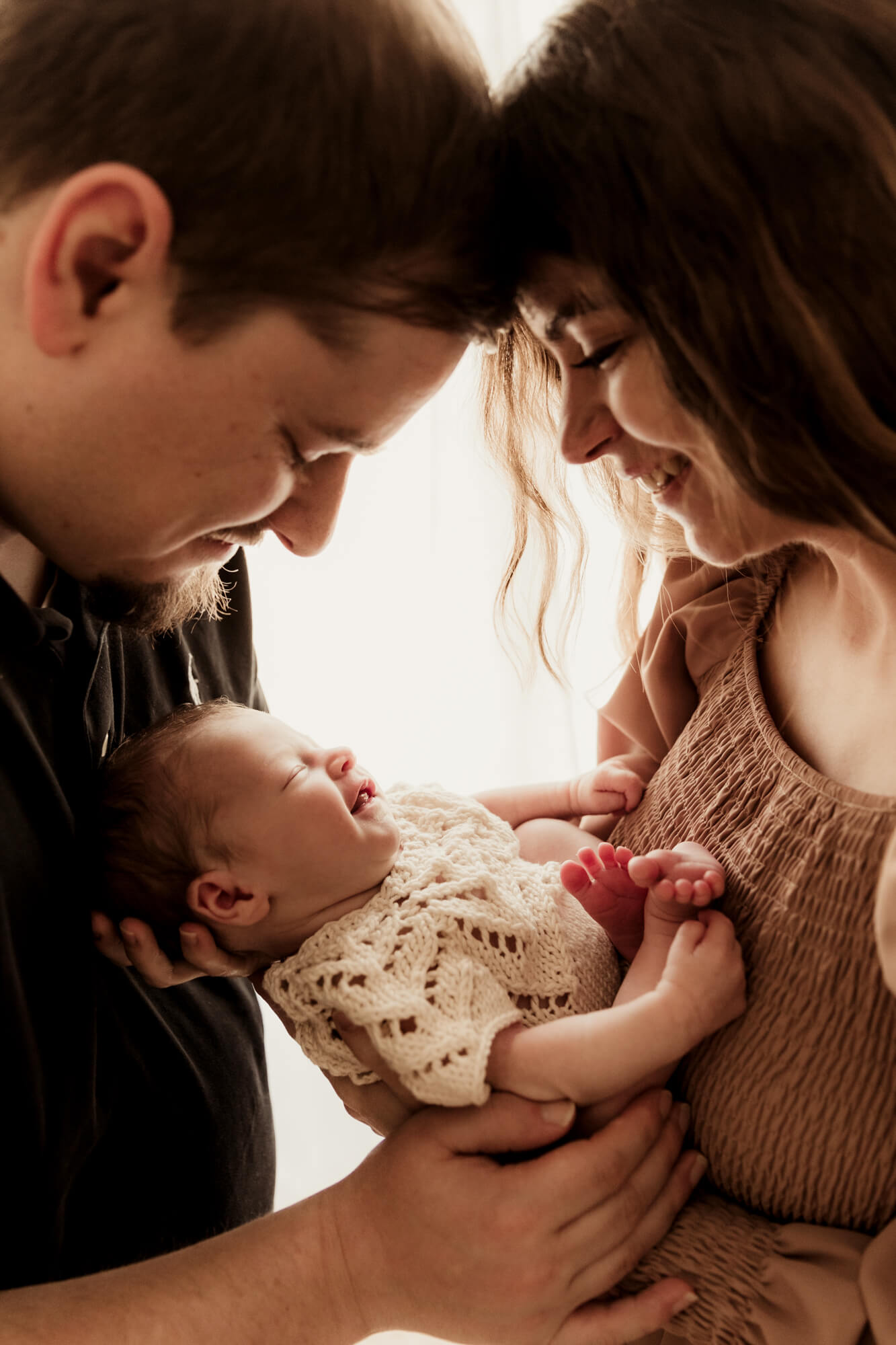 Happy new parents touch foreheads over their smiling newborn in between them in a window after some baby classes in OKC