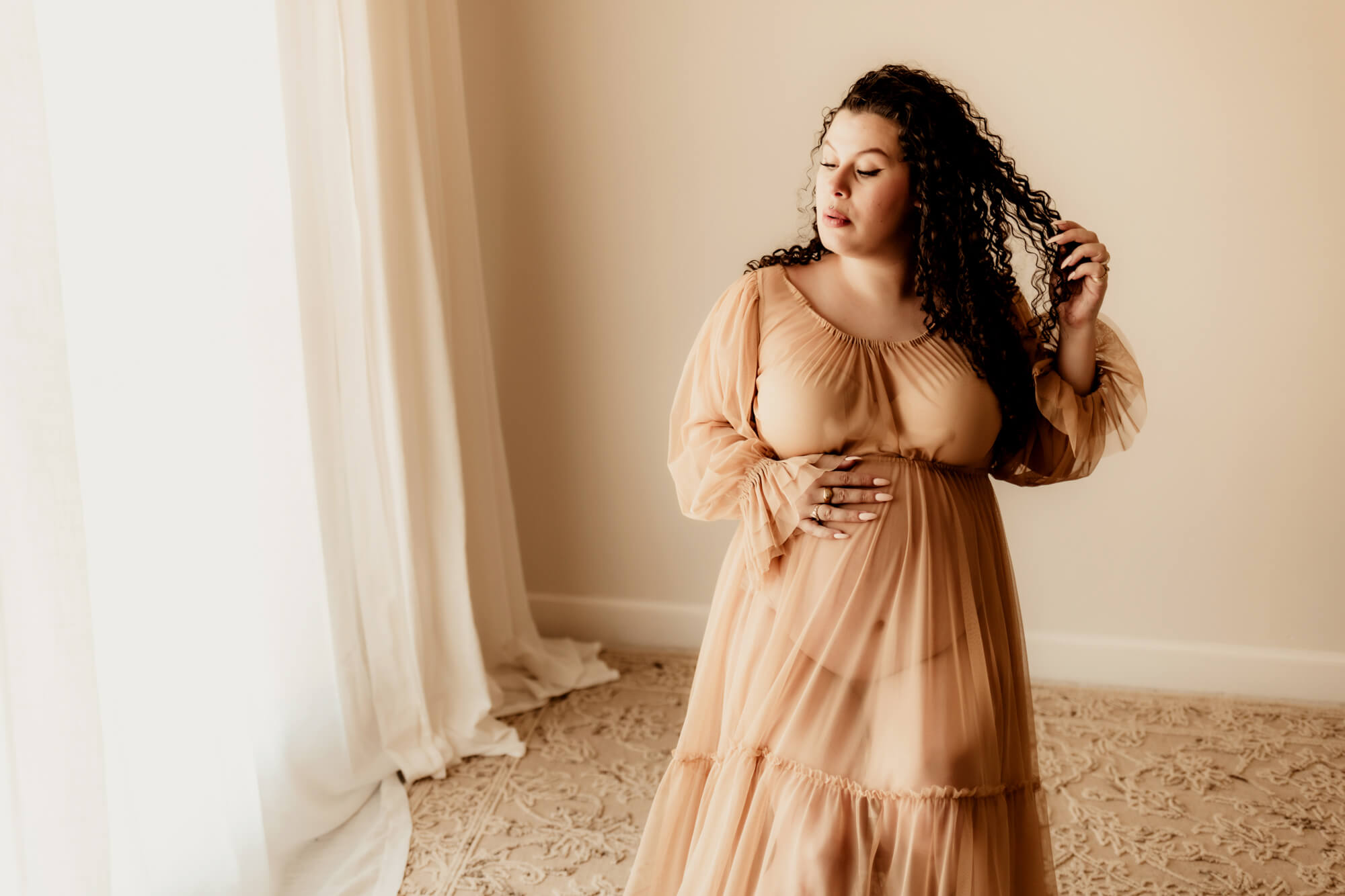 A mother to be rests a hand on her bump and plays with her hair in a studio