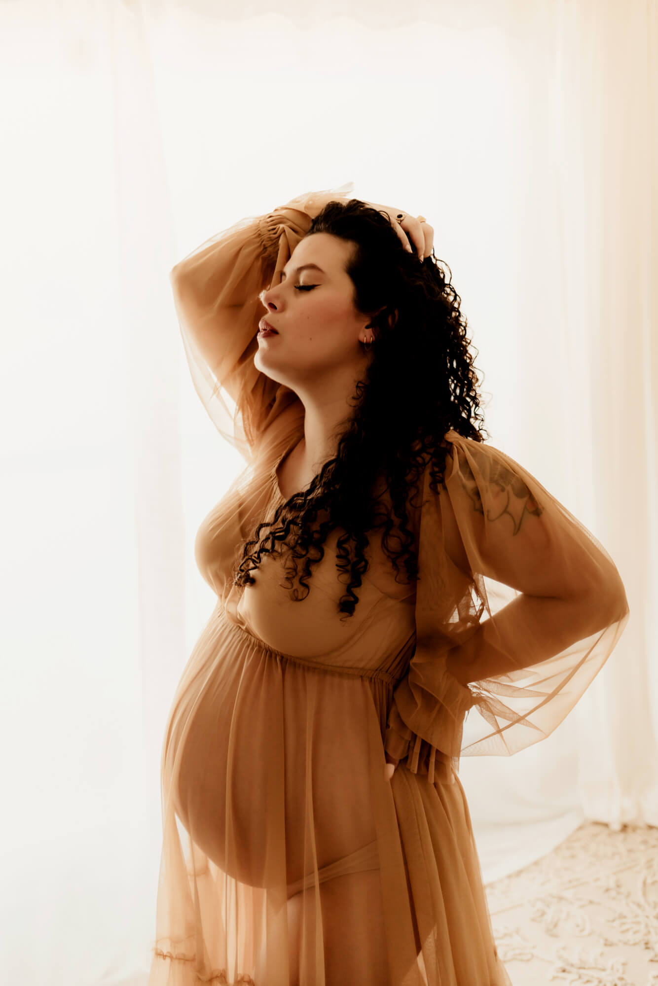 A pregnant woman holds her hair back and rests a hand on her back while standing in a window after some birthing classes in OKC