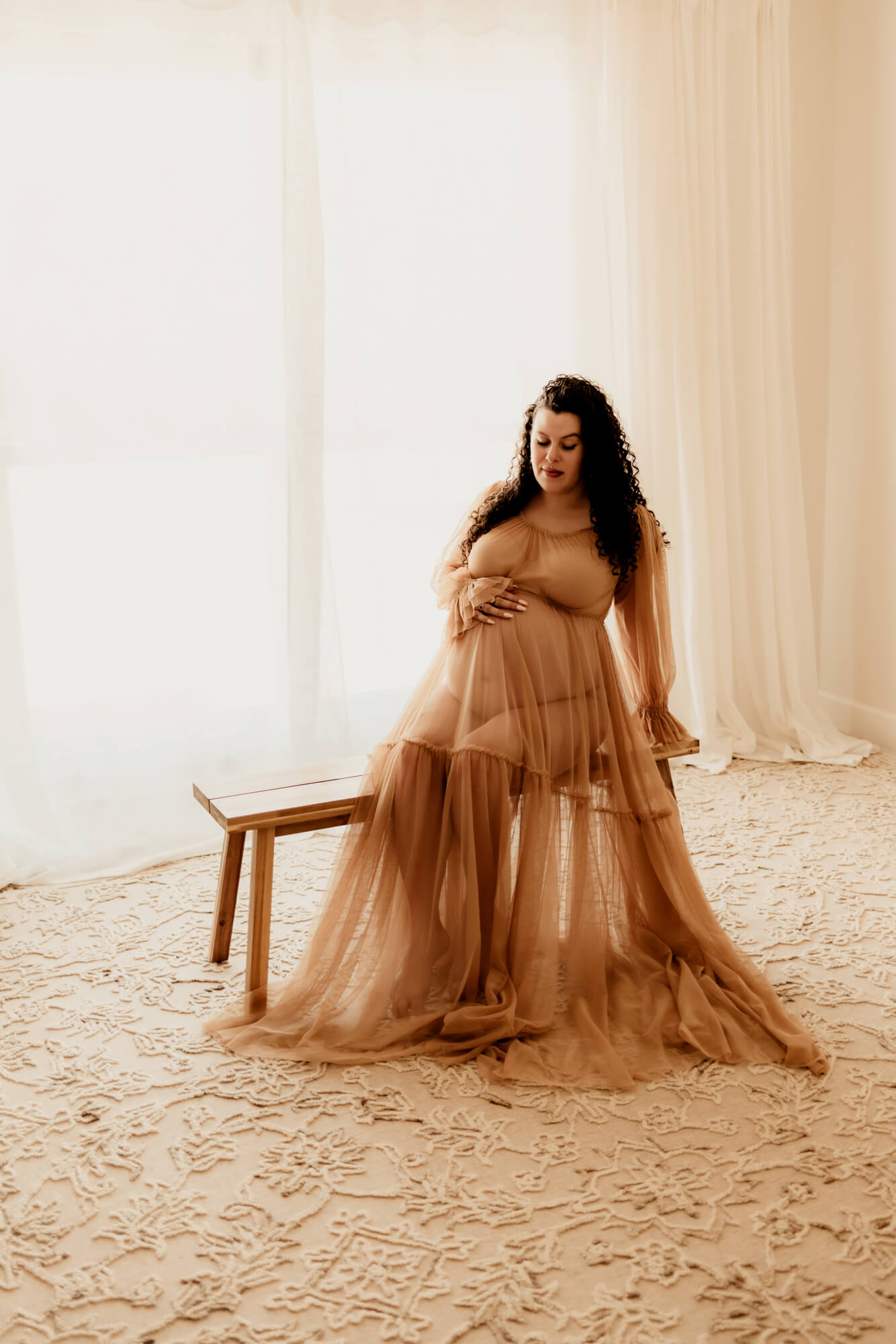 A mom to be leans back on a bench with a hand on her bump in a sheer maternity gown after some birthing classes in OKC