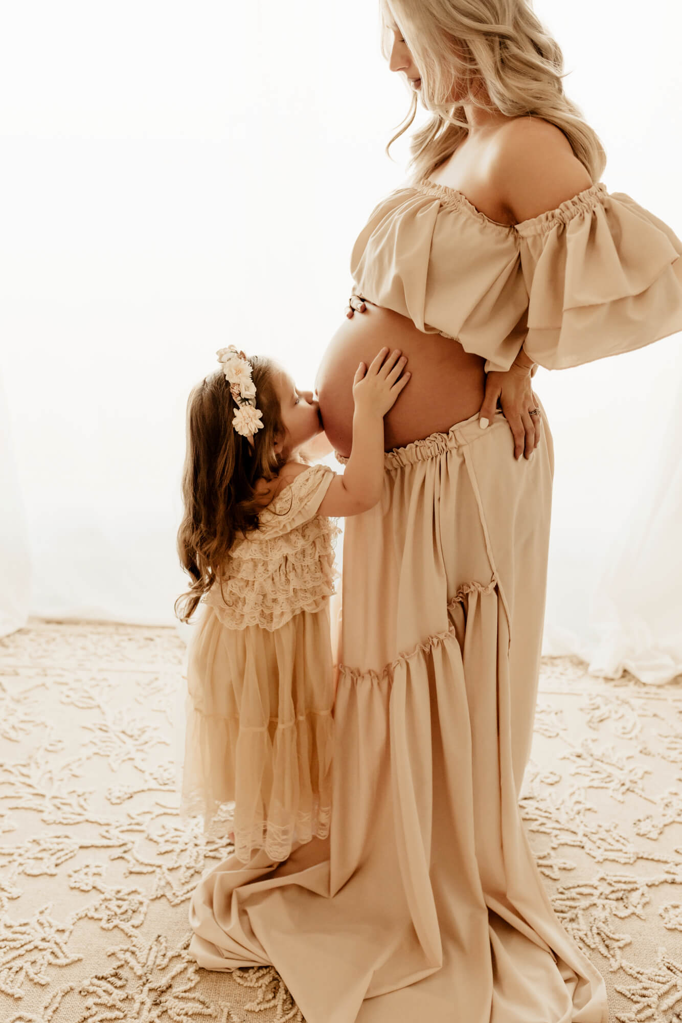 A toddler girl in a lace dress kisses mom's pregnant belly while standing in a studio after visiting Labor Of Love Midwifery