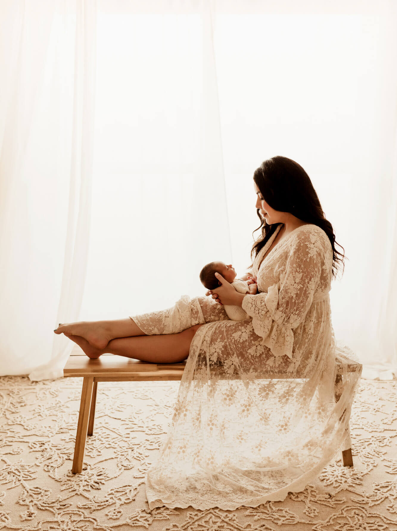 A new mother in a lace down sits across a wooden bench in a studio holding her newborn in her lap after getting a newborn screening in Oklahoma
