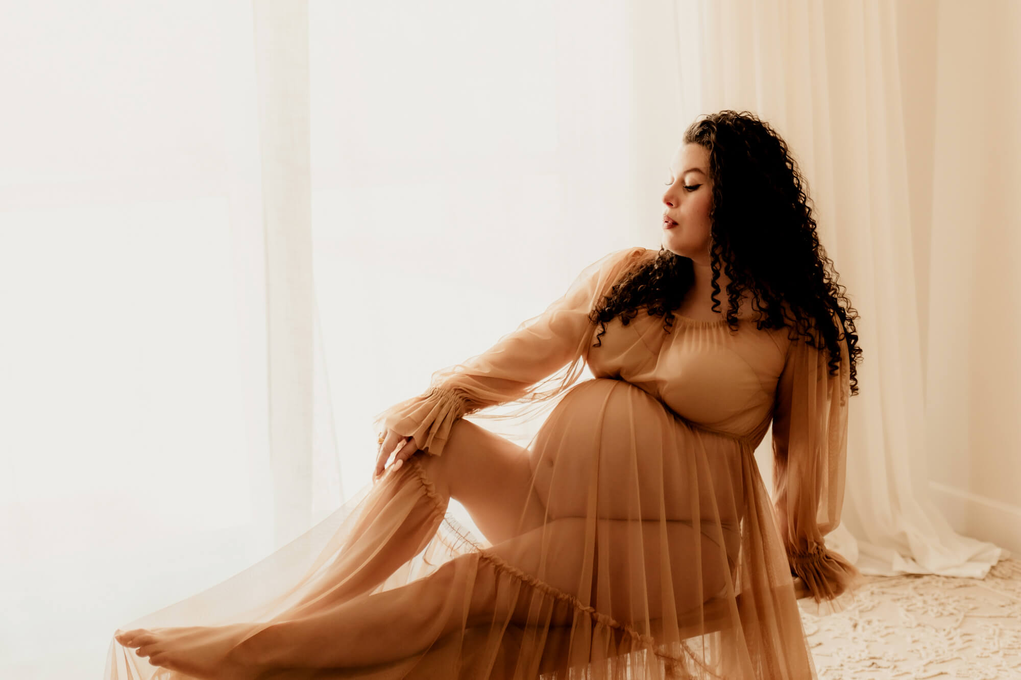 A proud pregnant woman sits across a wooden bench in a studio with a knee up