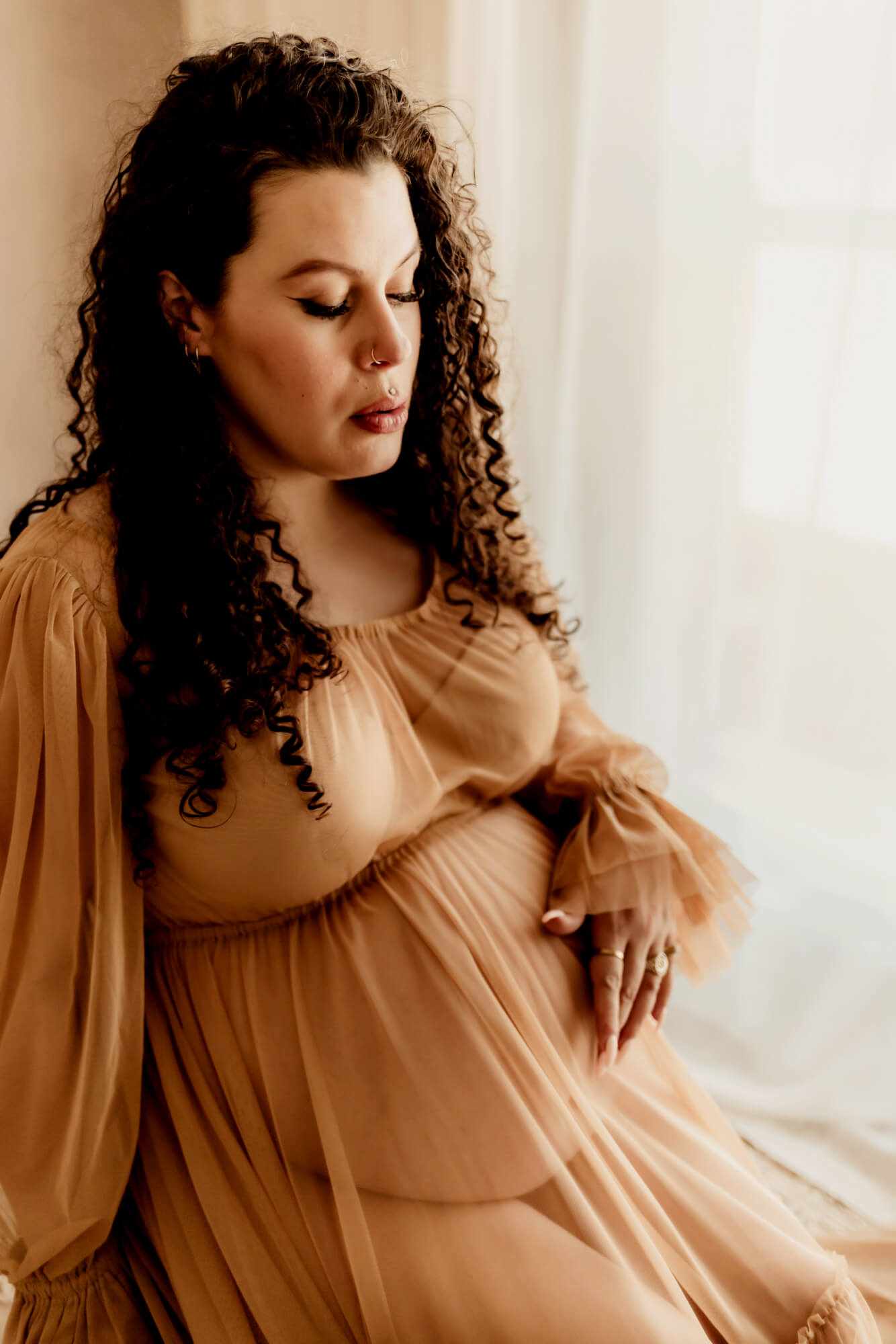 A pregnant woman sits on a stool in a window in a sheer maternity dress with a hand touching her bump after an OKC 4D Ultrasound