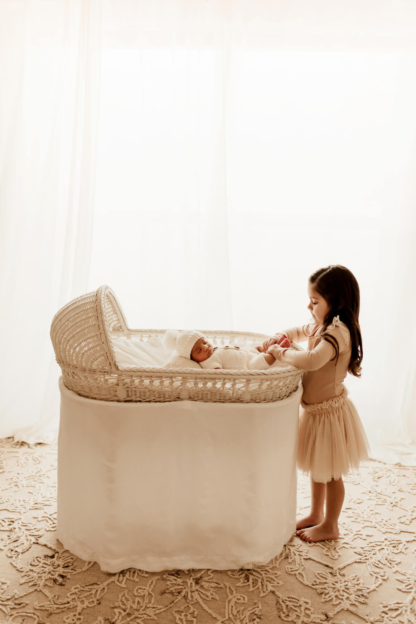 A toddler girl in a dress plays with the feet of her sleeping newborn sibling in a wicker crib