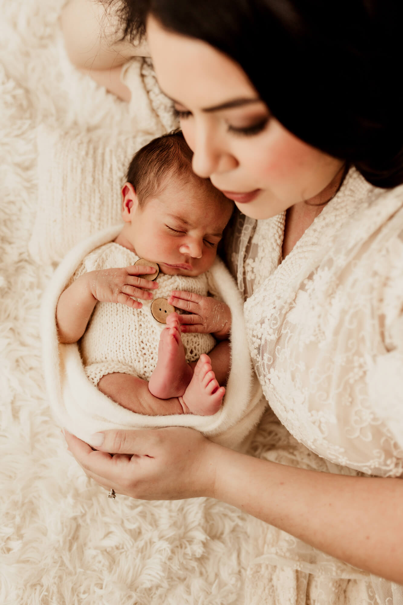 A new mom in a lace dress snuggles her sleeping newborn on a bed in a knit onesie