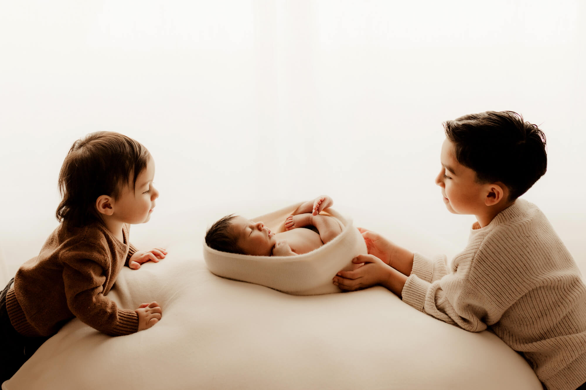 Older brothers look at their newborn baby brother sleeping in a wrapped swaddle.