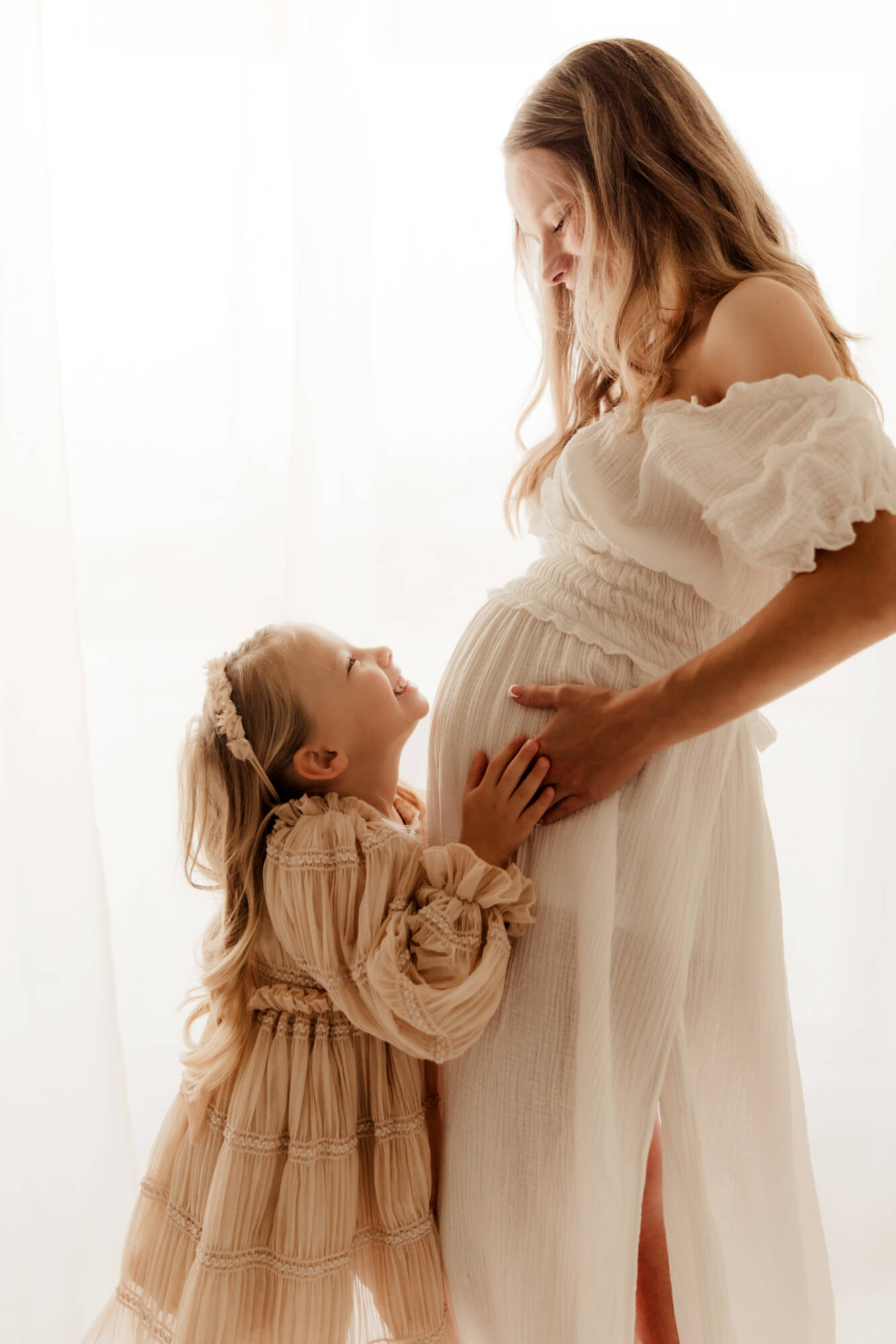 Girl wearing a pink dress smiles up at her pregnant mother.