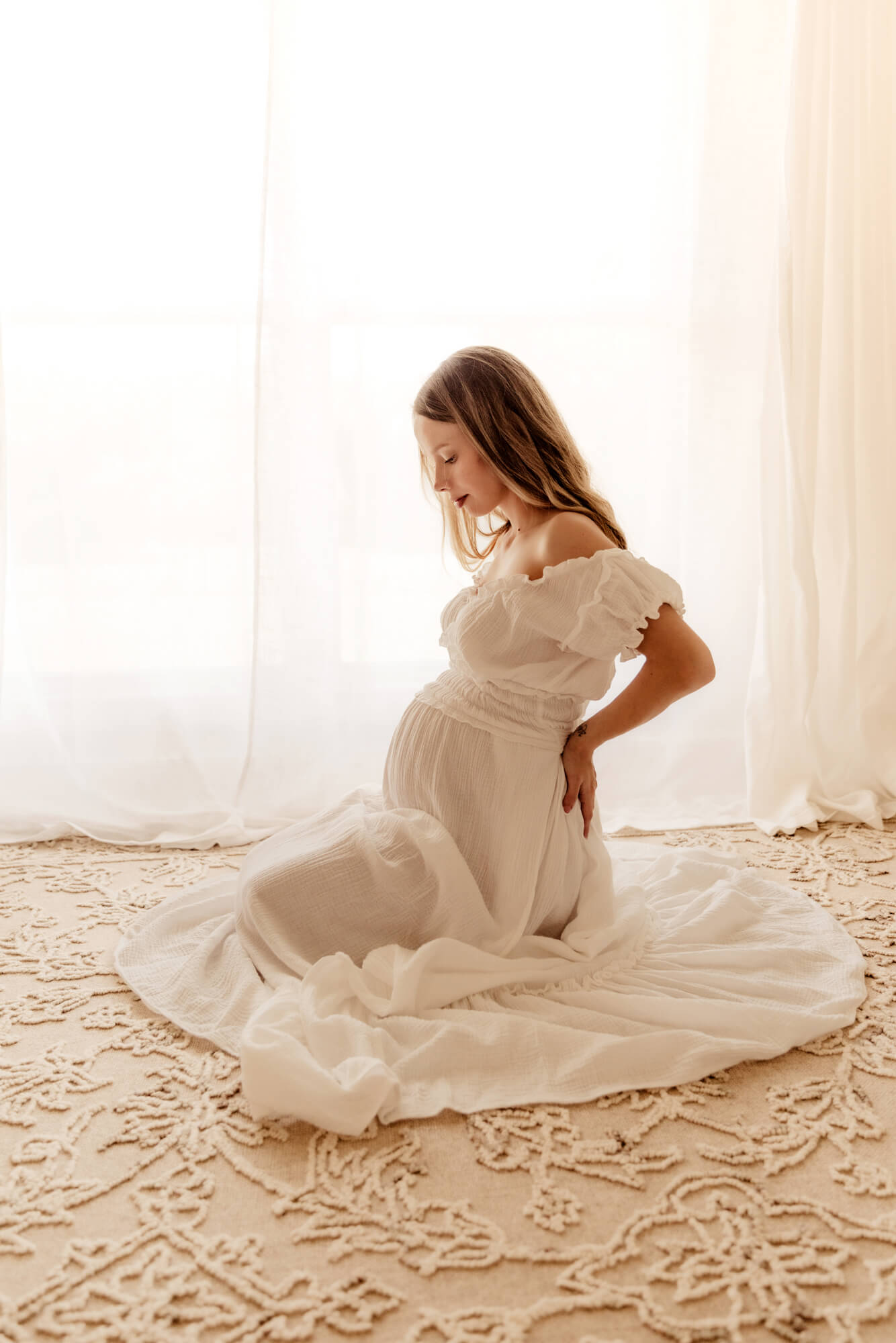 Pregnant woman with a white dress sitting a rug and looking at her belly, OBGYN Oklahoma City OK.