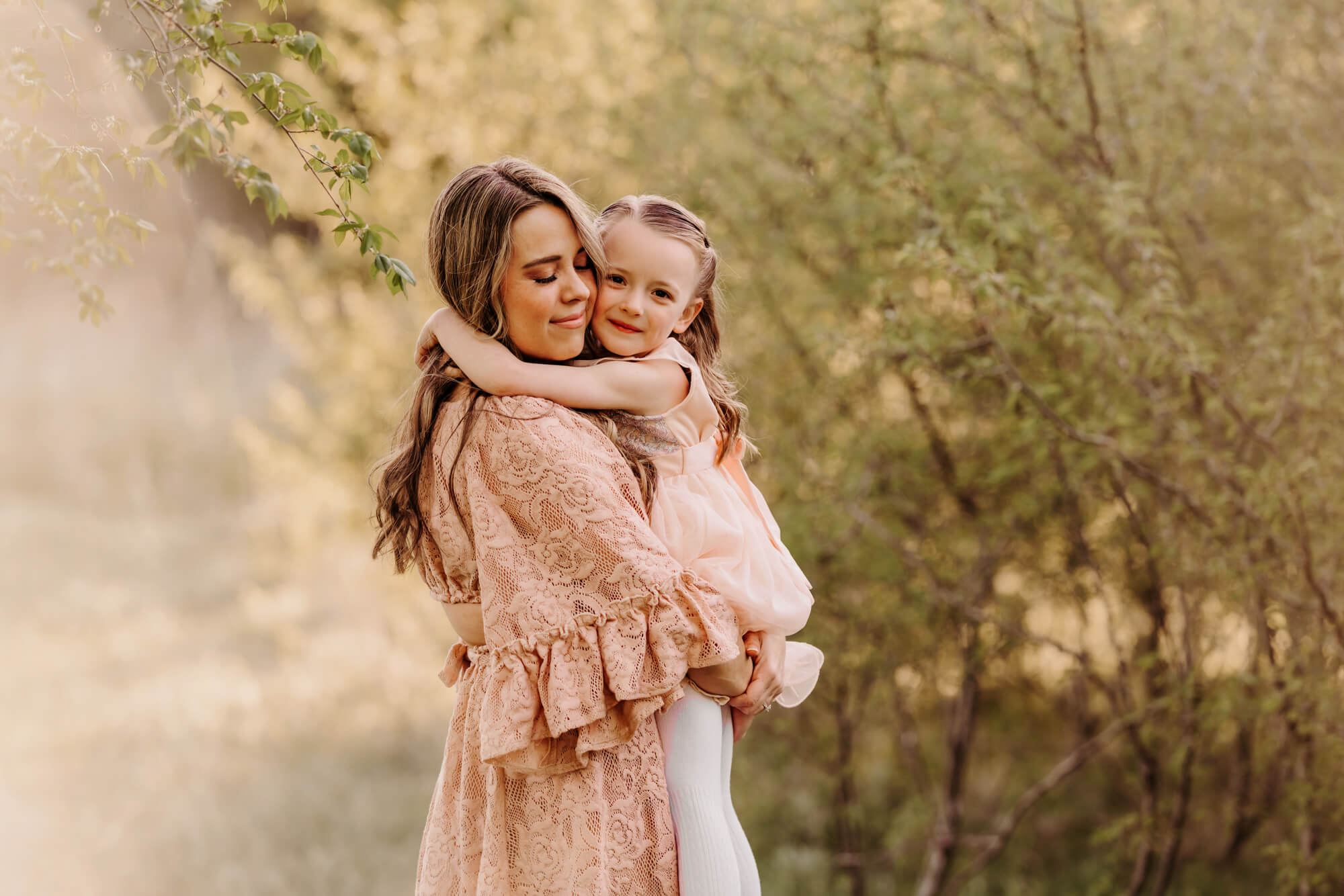 Mother wearing a pink lace dress smiles at her daughter with trees surrounding them, Montessori schools in OKC.