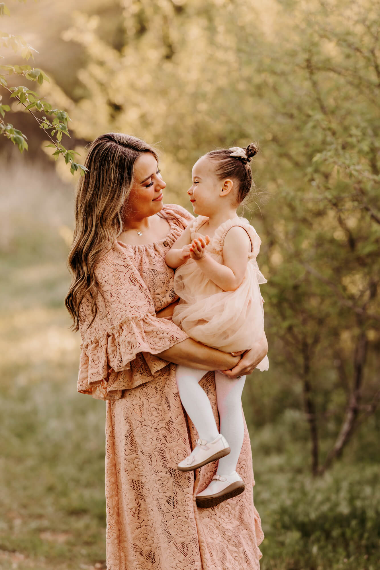 Mother wearing a pink lace dress smiles at her daughter with trees surrounding them, Montessori schools in OKC.