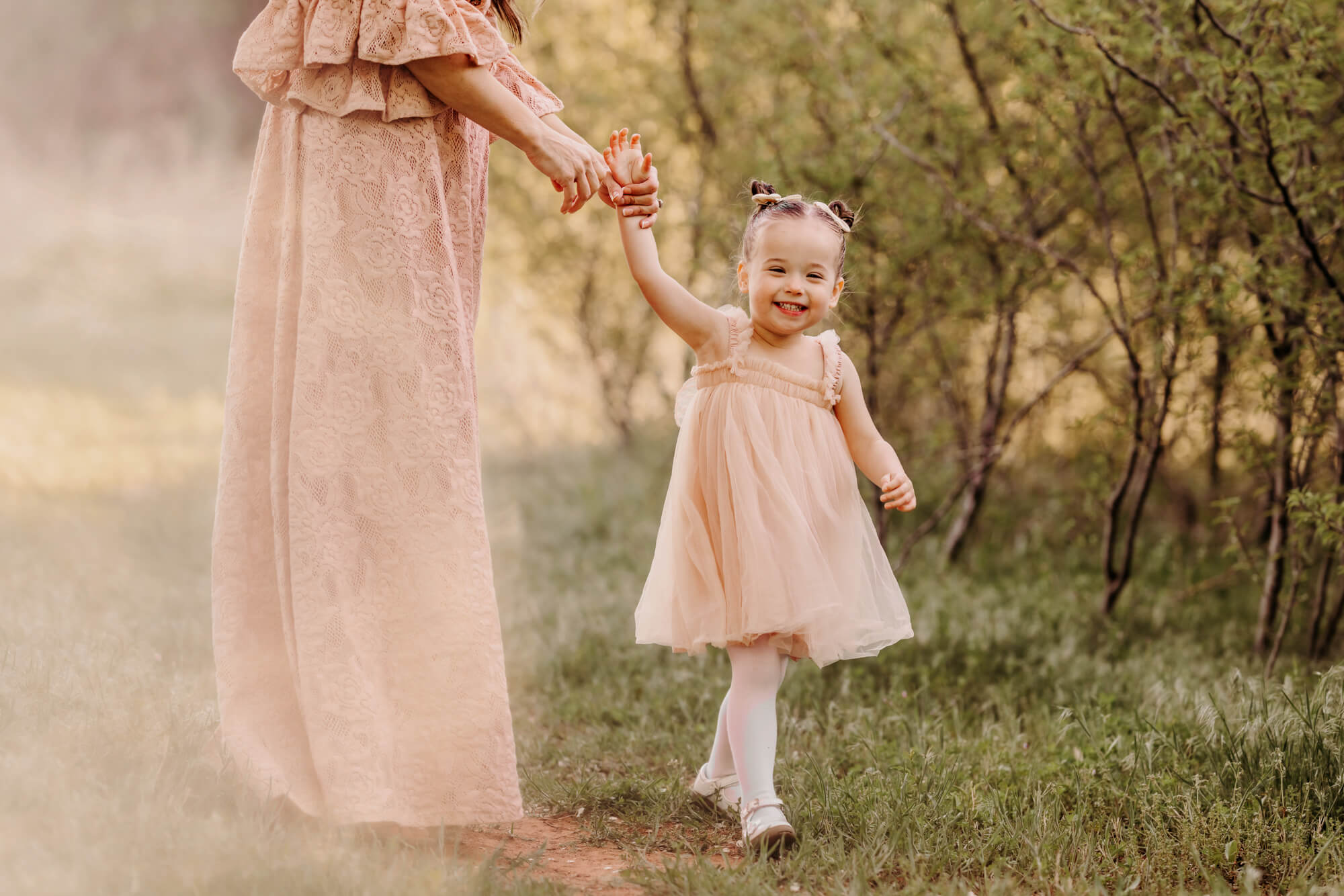 Girl wearing a pink dress smiles while holding her mother's hand, Mother wearing a pink lace dress smiles at her daughter with trees surrounding them, Montessori schools in OKC.