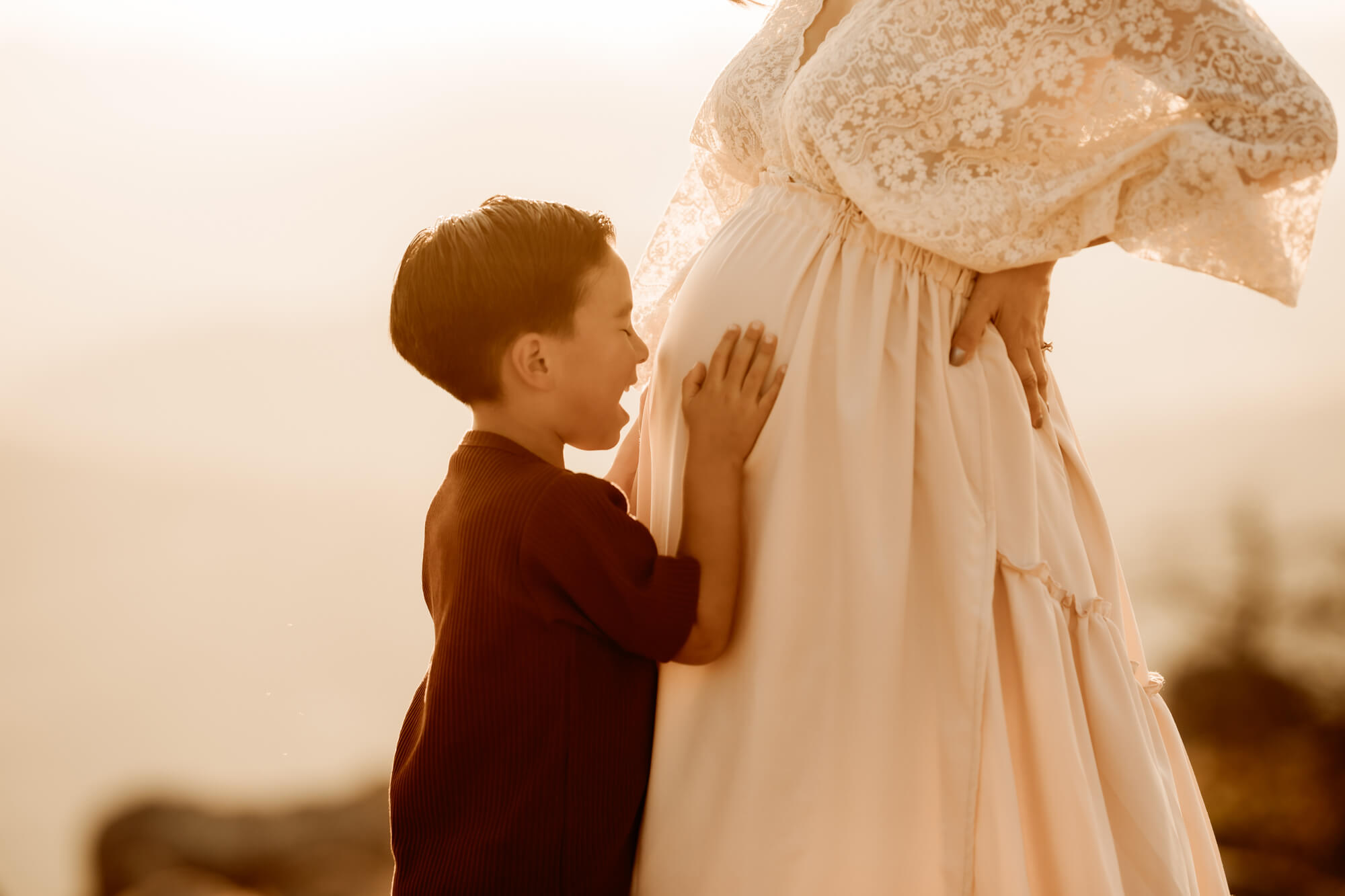 Boy smiles at his mother's belly while holding it with his hands.