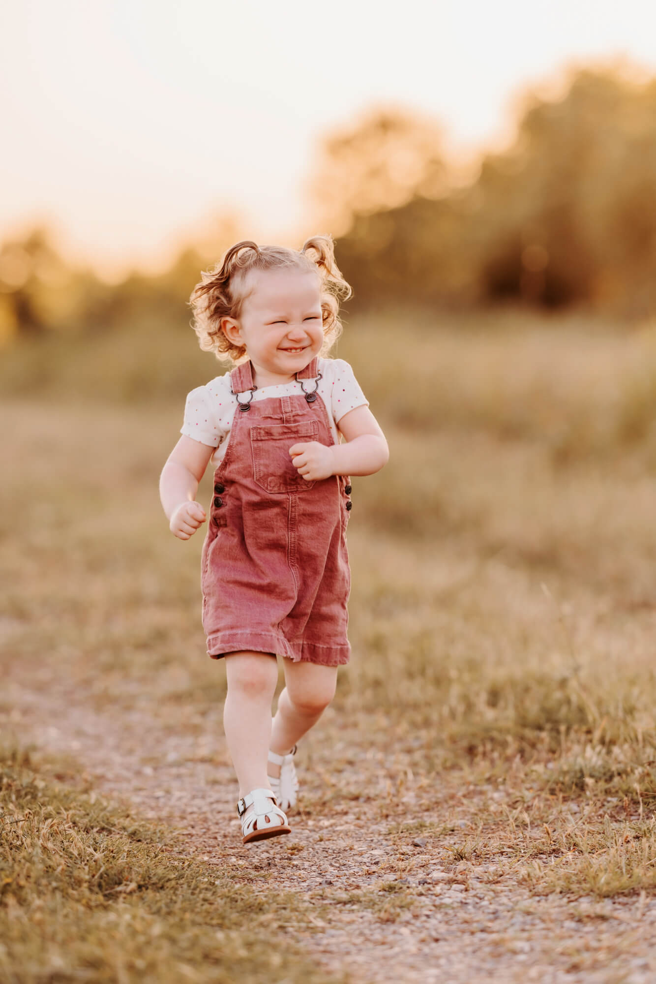 Girl wearing pink overalls running on a grassy road, OKC splash pads. 