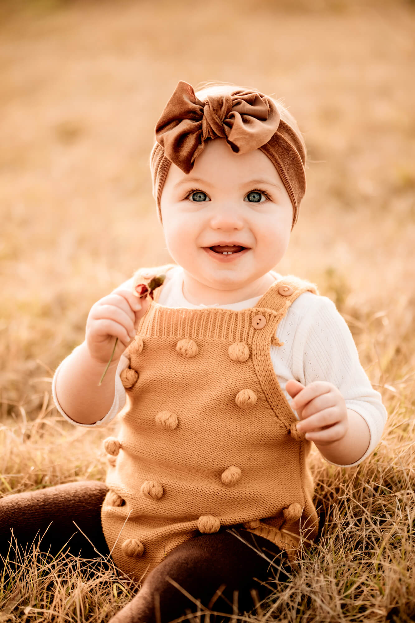 Girl with a yellow romper and brown bow smiling while sitting on the grass.