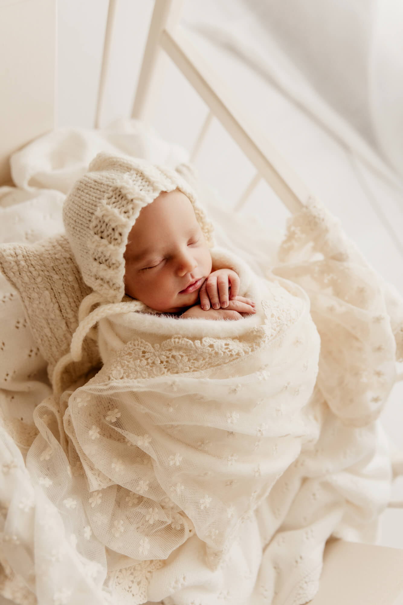 Baby girl wearing a white bonnet and swaddled is sleeping on a small pillow in a crib.