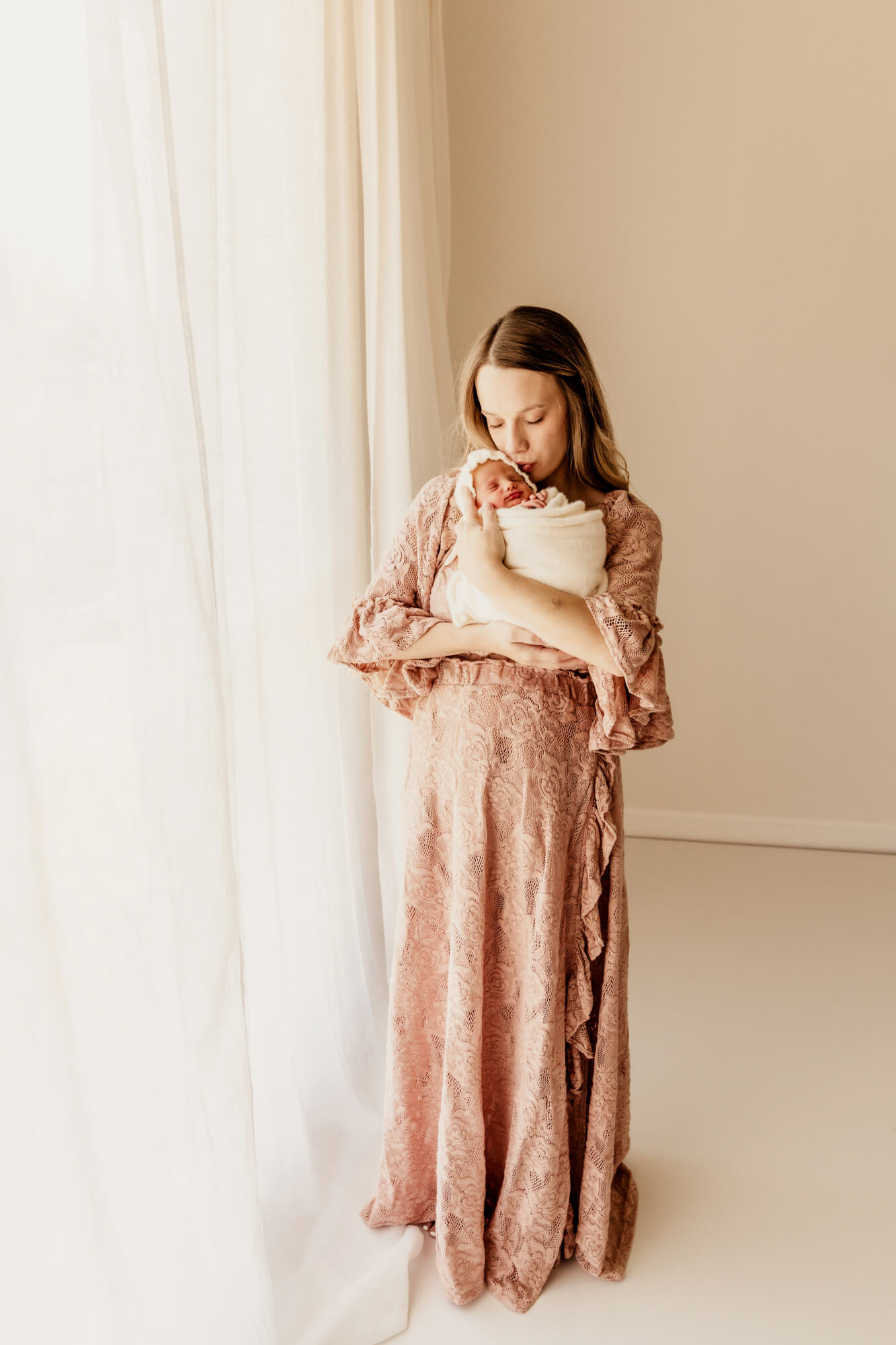 Mother kisses her baby girl while standing next to a window, postpartum doula OKC.