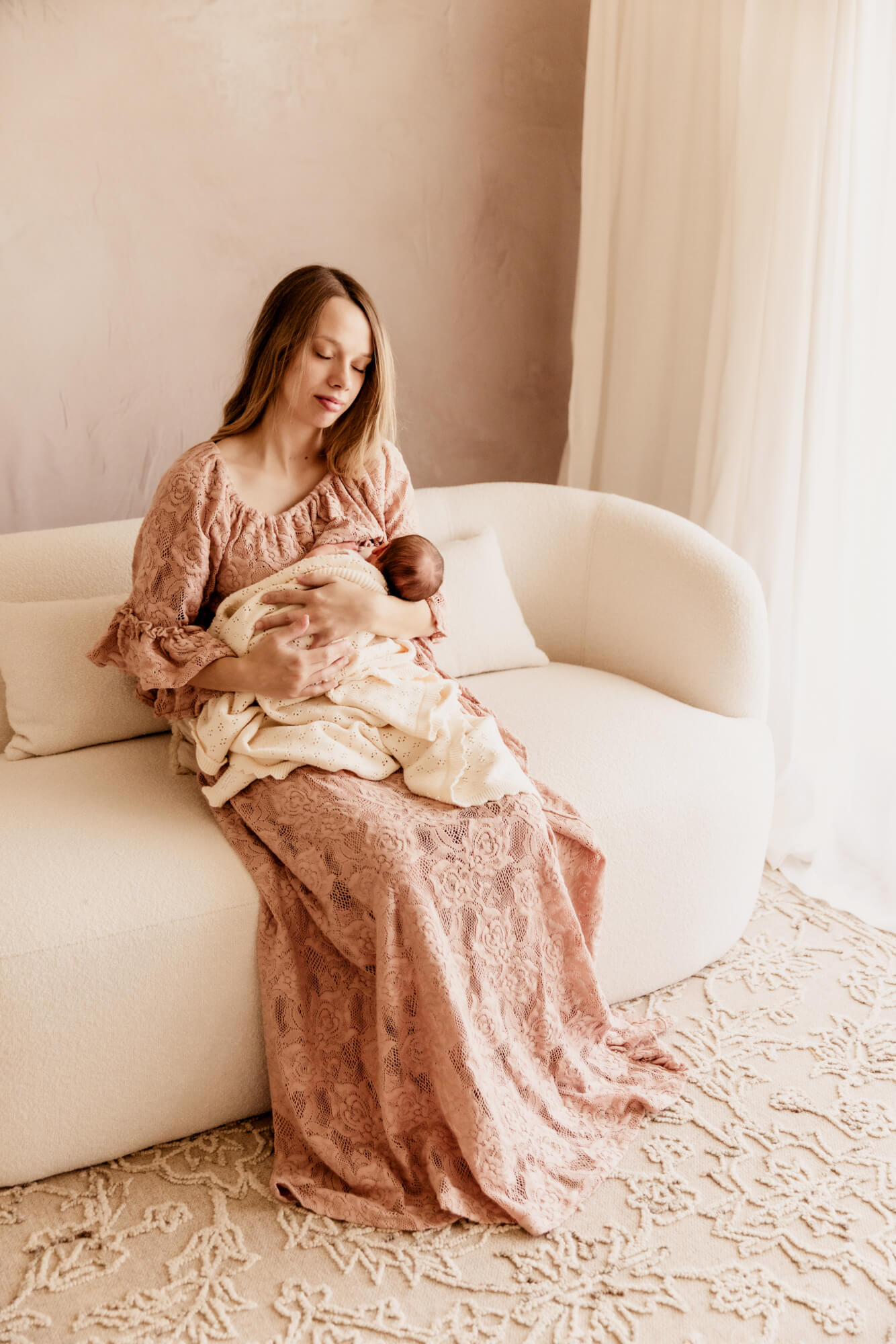 Postpartum mother nurses her baby while sitting on a white couch, postpartum doula OKC.
