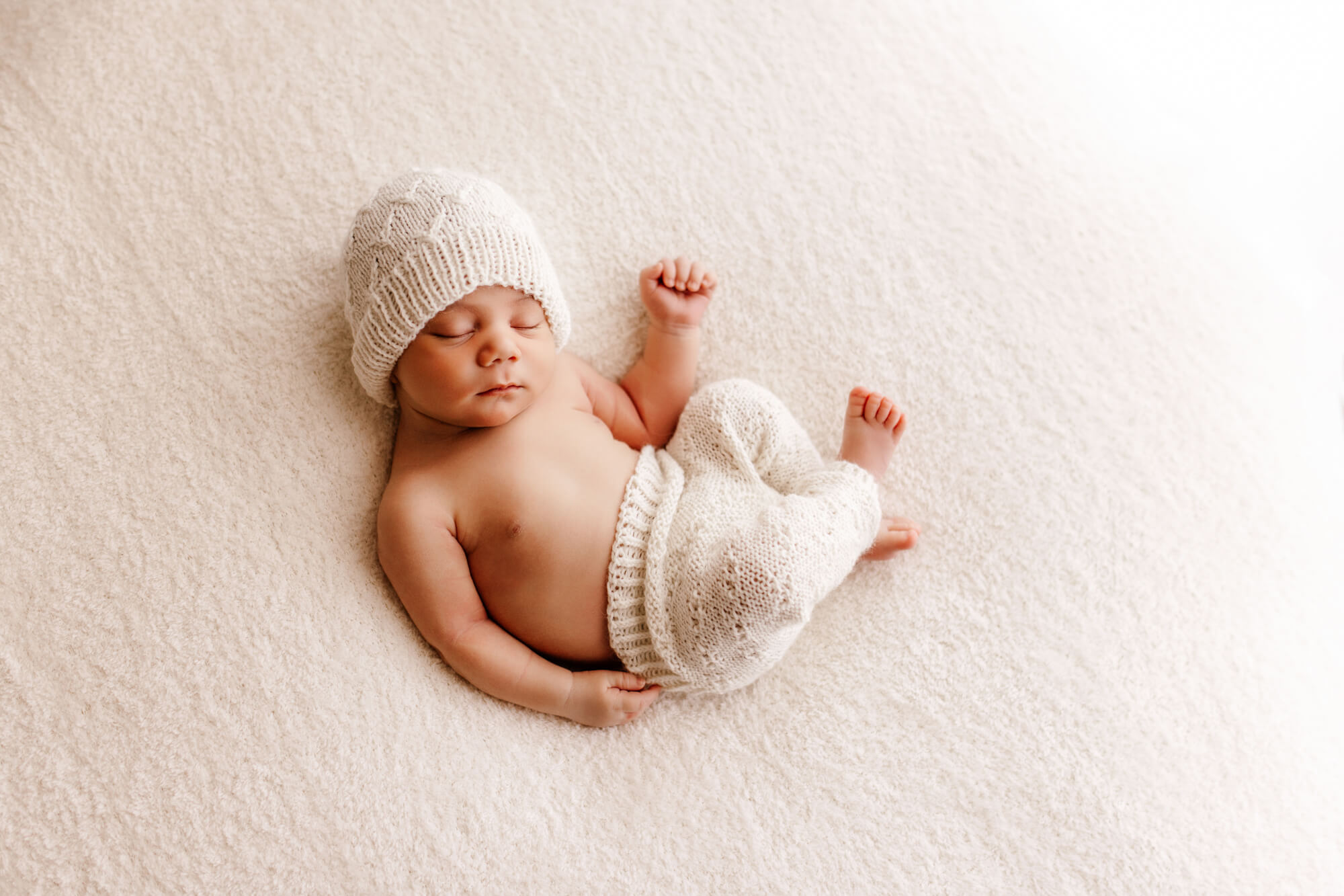 A newborn baby lays in knit pants and matching beanie on a bed after enjoying infant swim lessons in OKC
