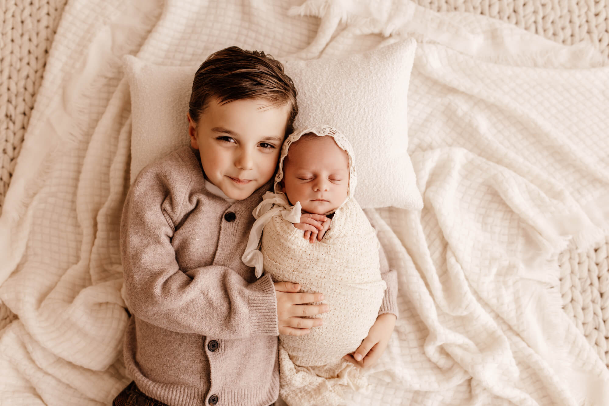 A toddler boy lays smiling on a bed cuddling his sleeping newborn abby sister
