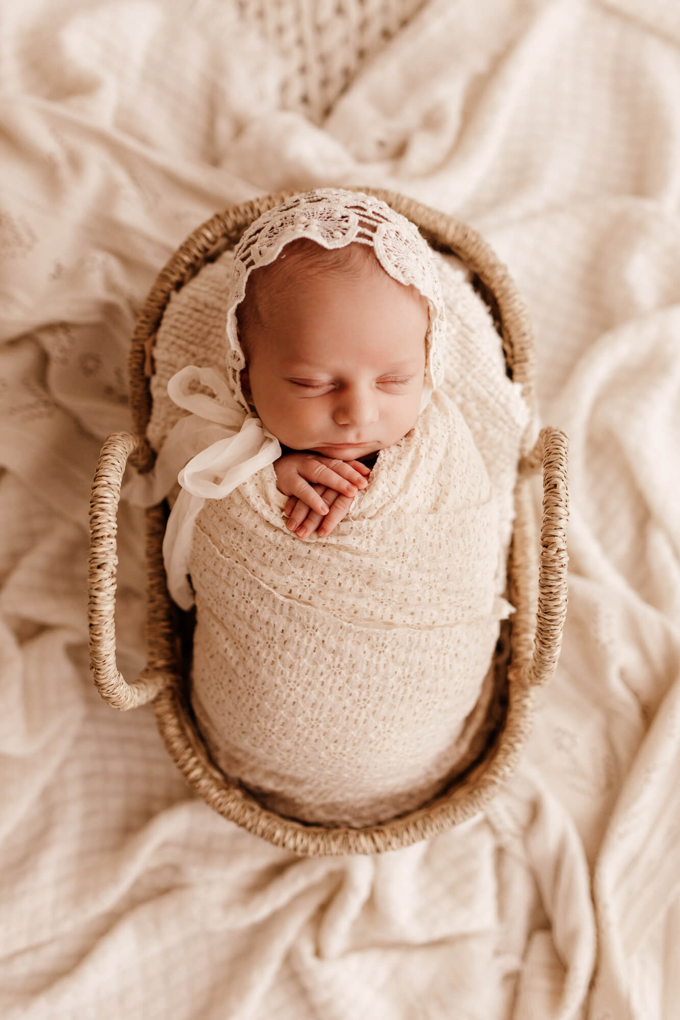 A newborn baby girl sleeps in a white swaddle and lace bonnet in a woven basket before searching for Norman, OK daycares