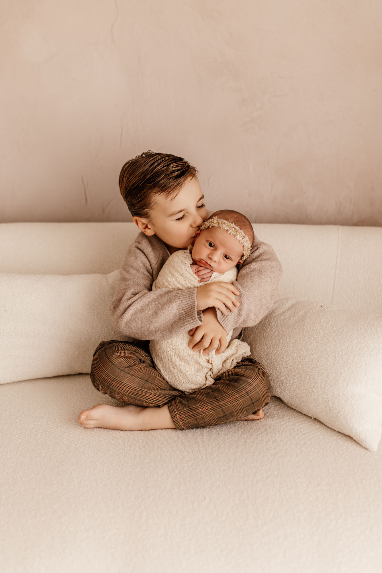 A toddler boy in a sweater kisses his newborn baby sister in his lap on a couch after finding Norman, OK daycares
