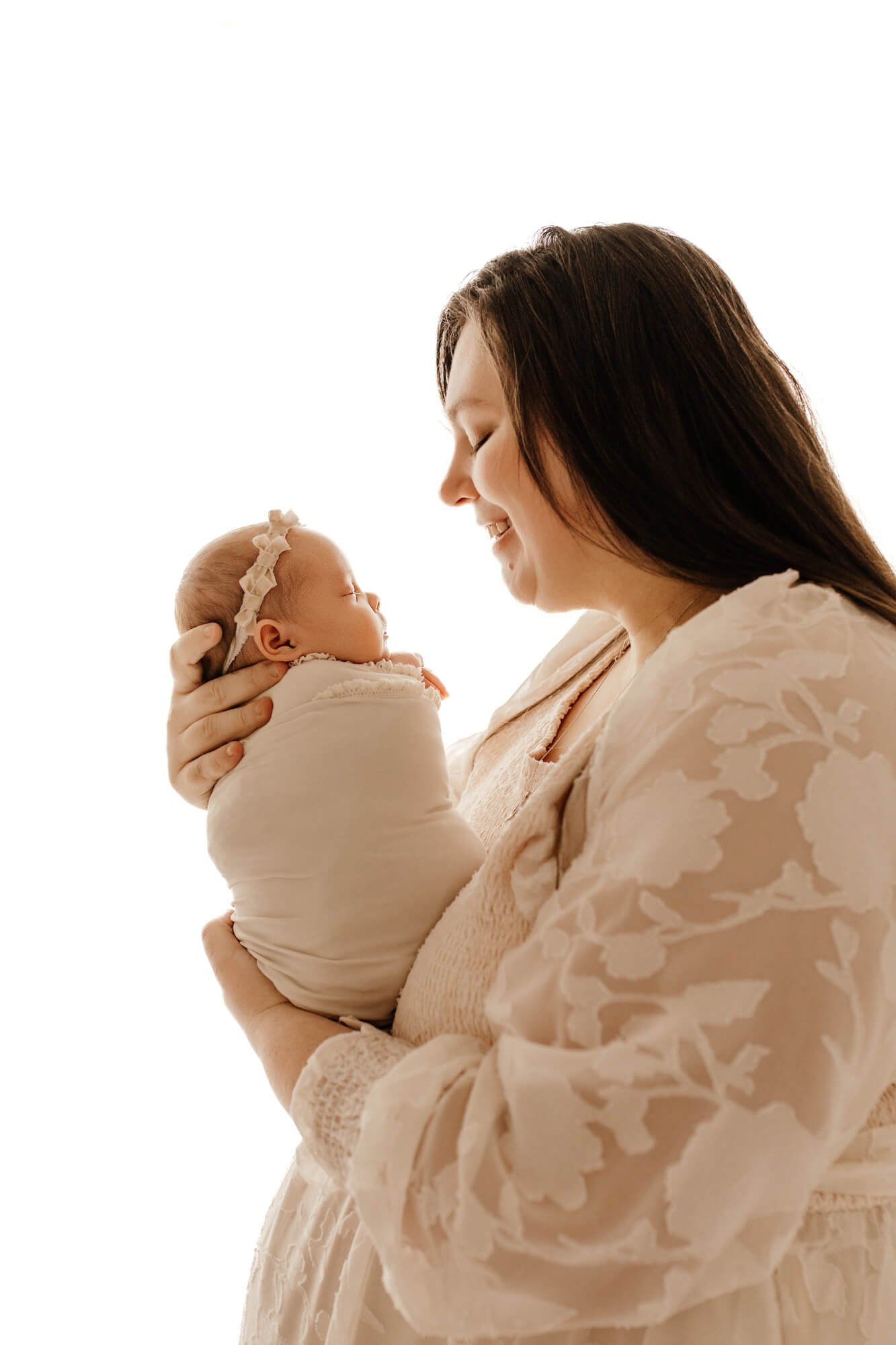 A happy new mom in a lace gown smiles at her sleeping newborn in her hands in a tight swaddle