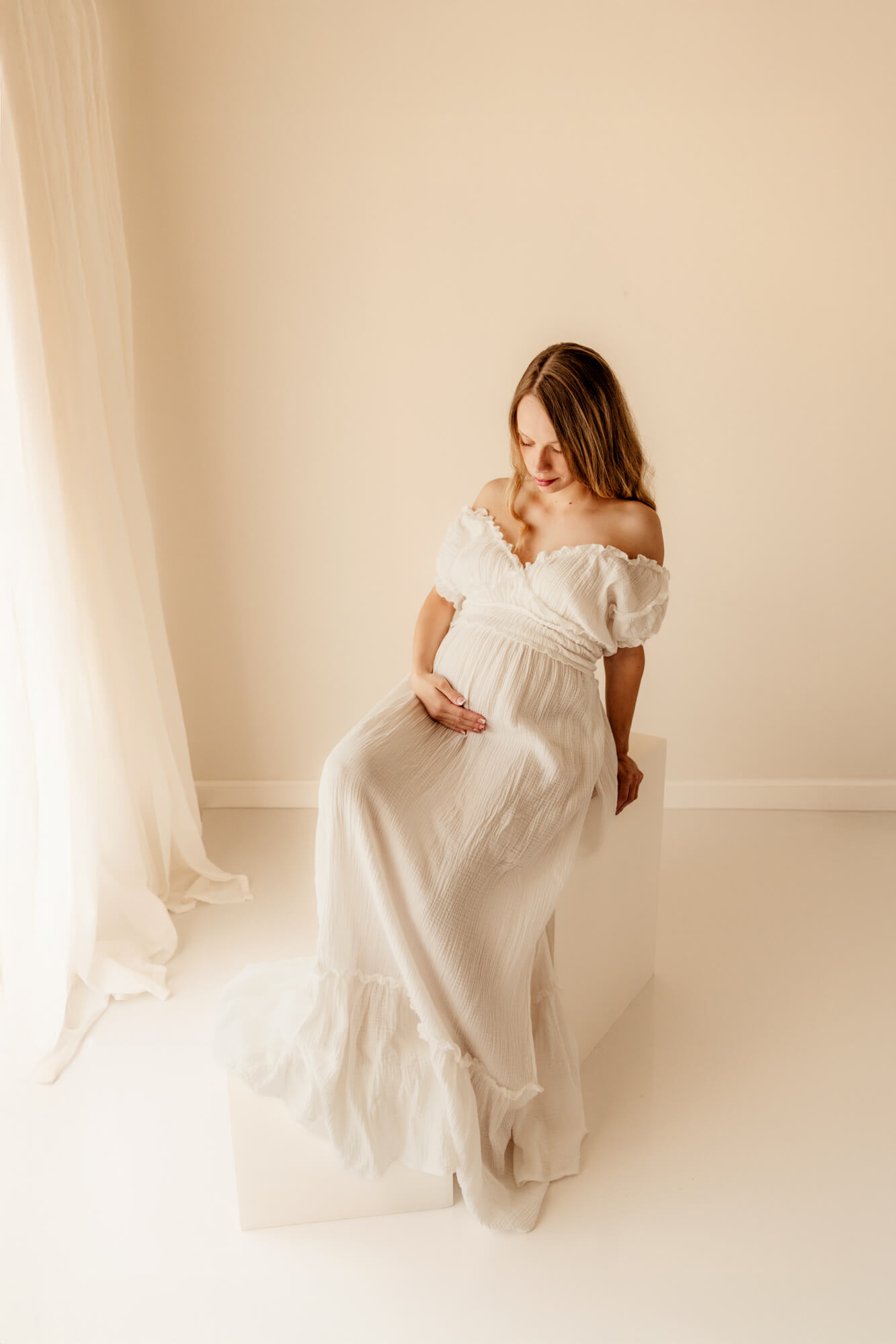 A happy mother to be sits on white boxes in a studio under a window in a white maternity gown gazing down to her bump after enjoying Anticipation Ultrasound Studio OKC