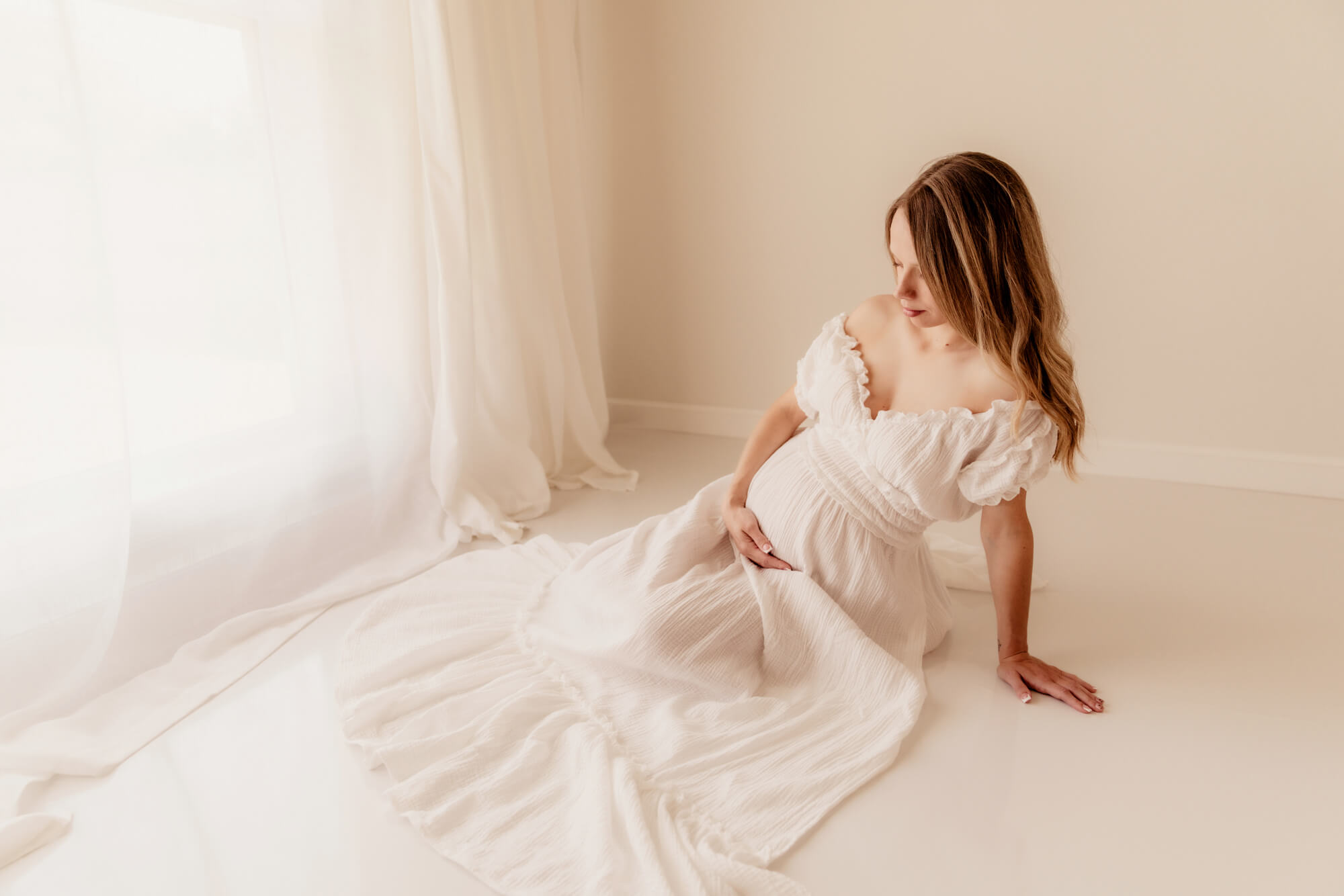 A mother to be sits under a window on the studio floor in a white maternity dress after visiting Anticipation Ultrasound Studio OKC