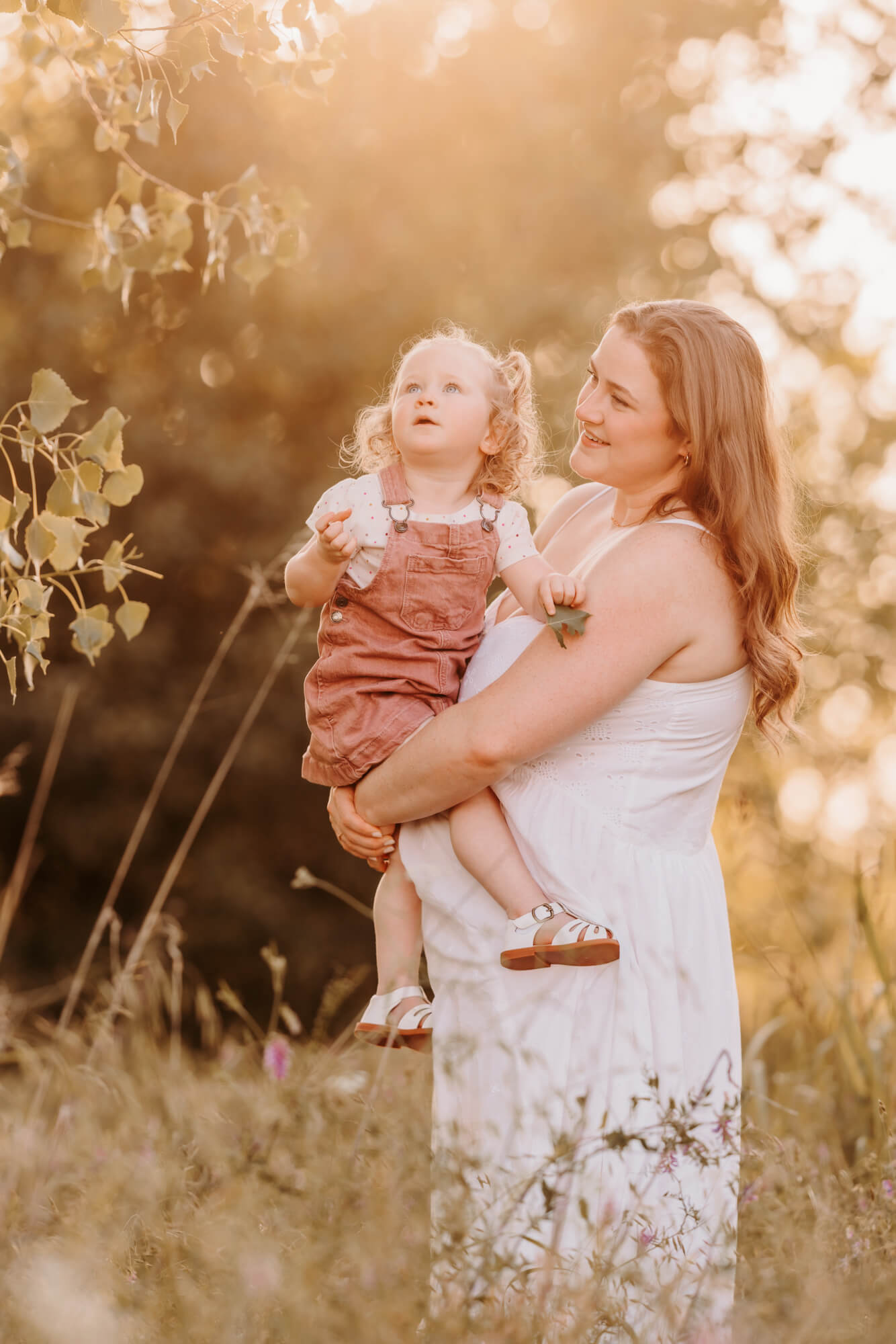 A curious toddler girl in mom's arms looks up to a leaves on a tree in a park at sunset