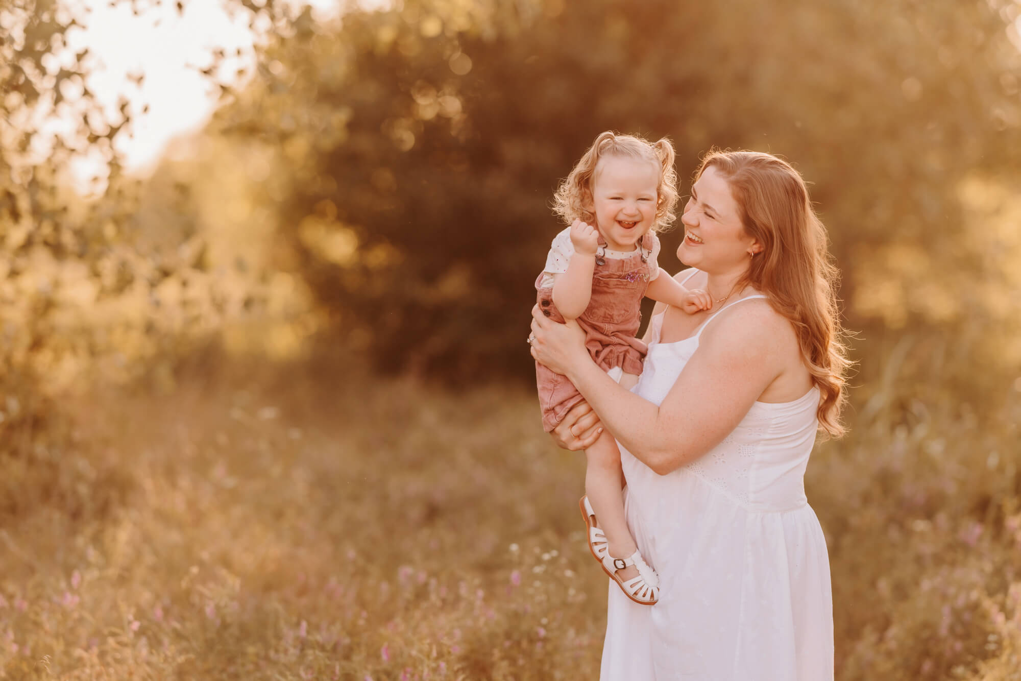 A happy mother in white laughs with her toddler daughter in pink overalls in a park at sunset after a Mother's Day Out in Edmond, OK