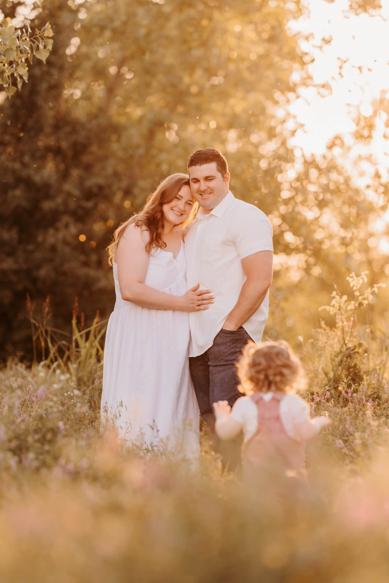 Happy mom and dad in white stands in a park as their toddler runs towards them at sunset after a Mother's Day Out in Edmond, OK